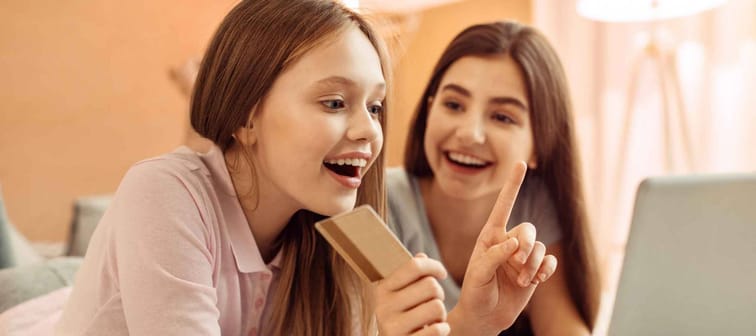 Incurable shopaholic. Excited pretty teenage girl lying on the bed next to her elder sister and choosing an item in an online shop while holding a golden credit card