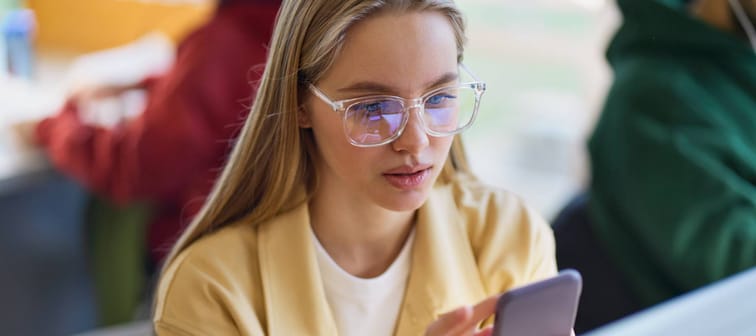 Young bespectacled blonde woman looking at her phone with some concern.
