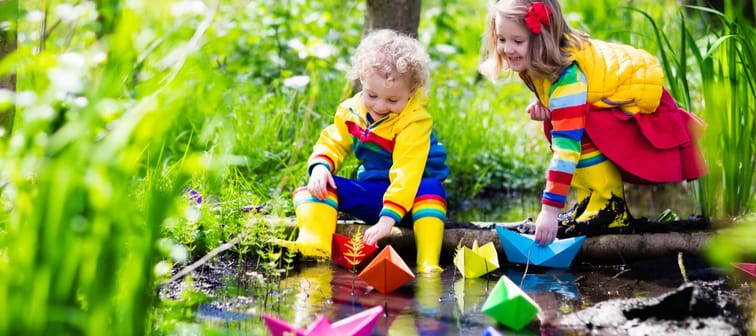 Children play with colorful paper boats in a small river on a sunny spring day.