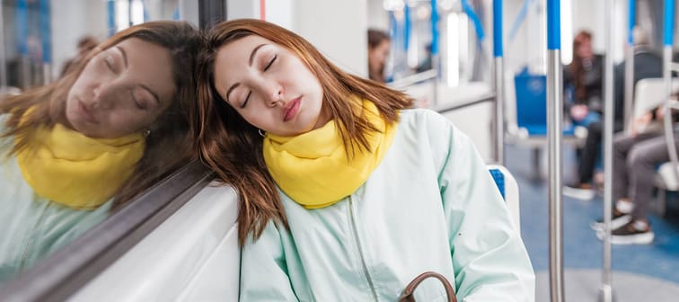 A brightly-dressed Gen Z female riding the subway asleep, and resting her head against the window.