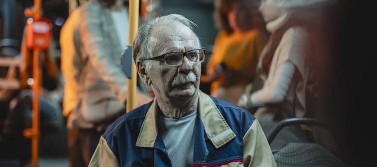 Older man with white hair and moustache riding public transit and looking deep in thought and unnerved.
