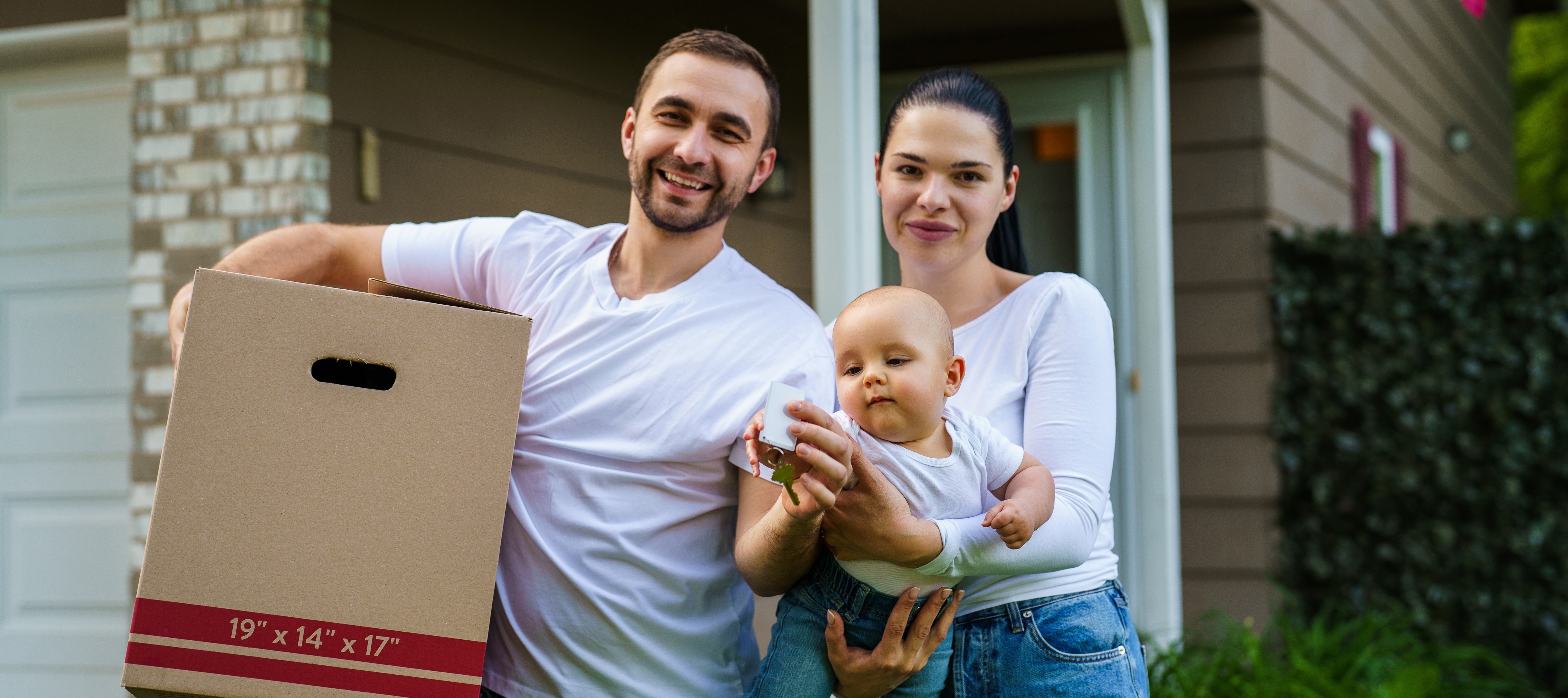Happy family carrying cardboard boxes with their belongings, smiling to the camera, posing in front of their new home.