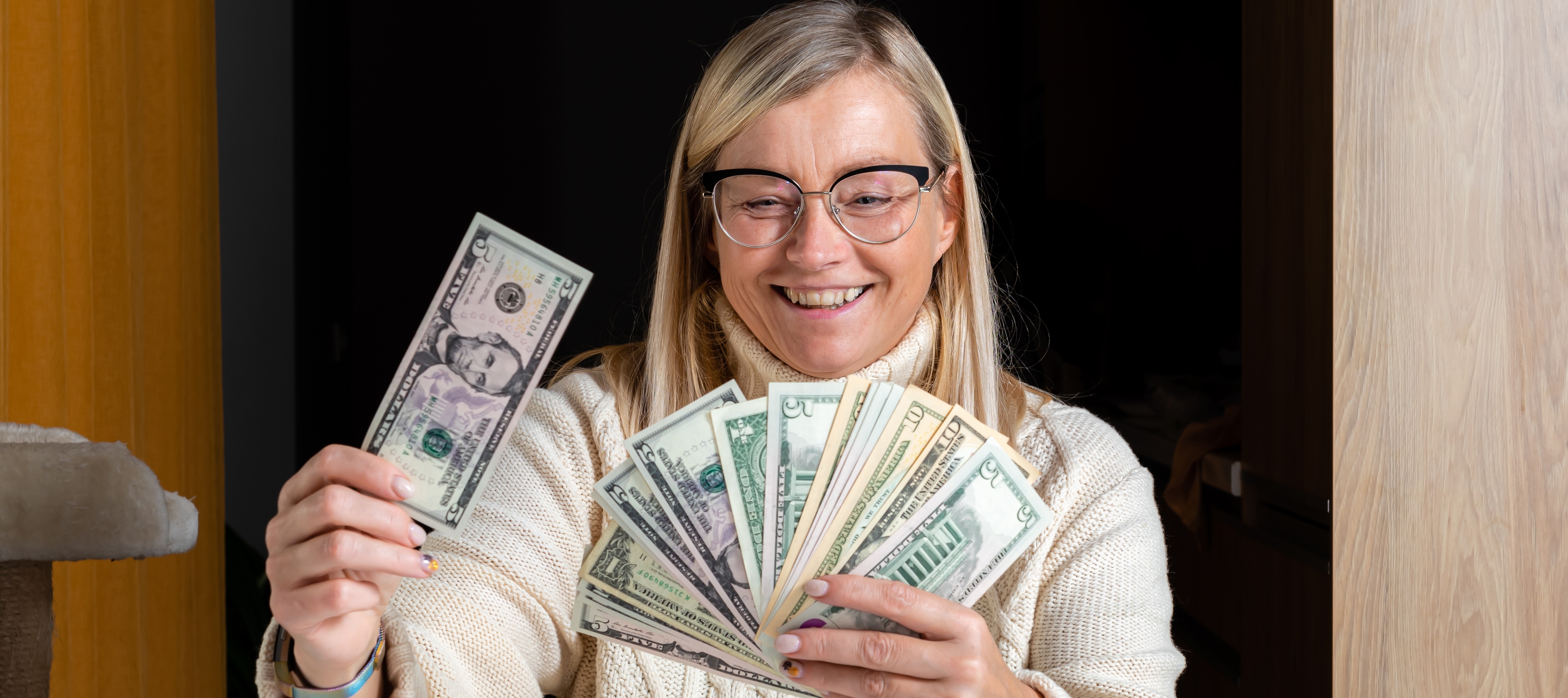 Happy woman sitting at a table and holding dollar bills with a journal and change on the table in front of her.