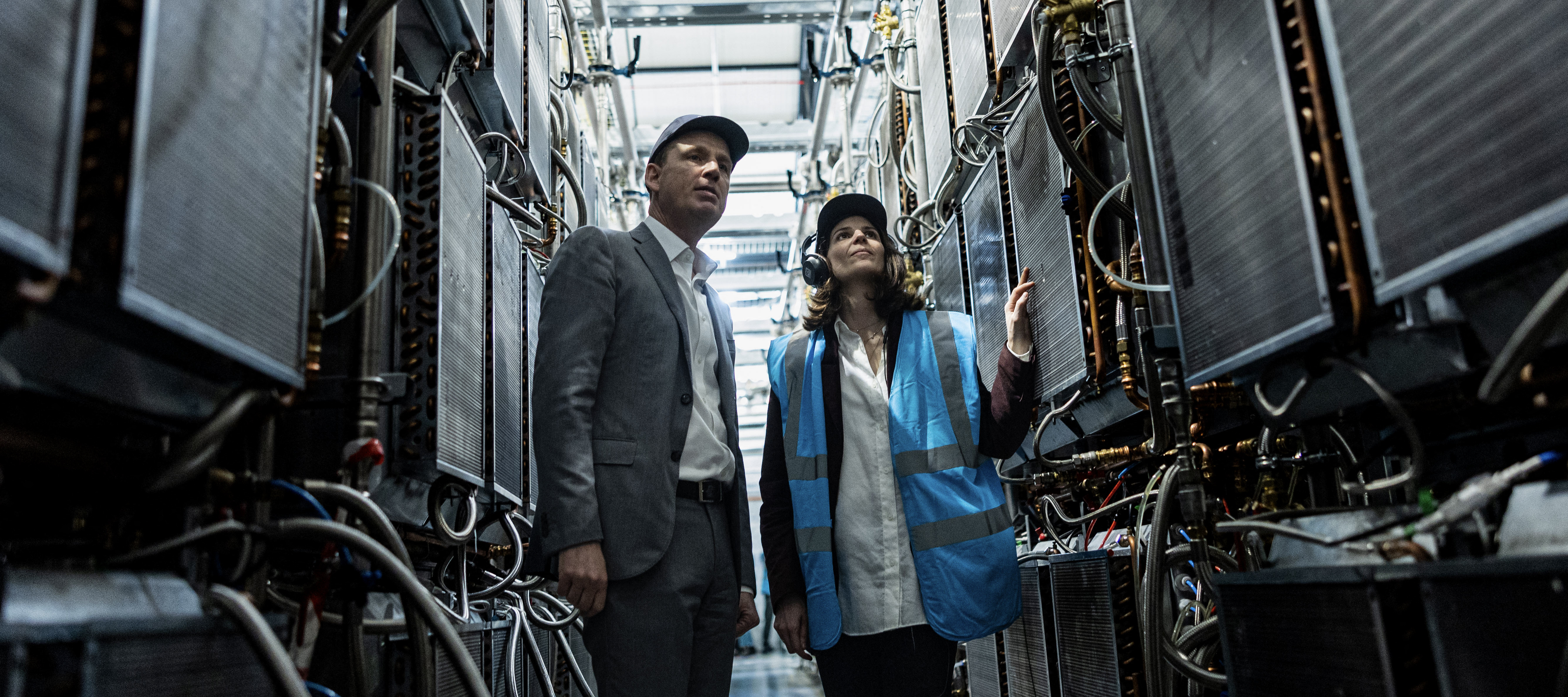Man and woman touring data center with power grids and cables surrounding them in narrow passageway.
