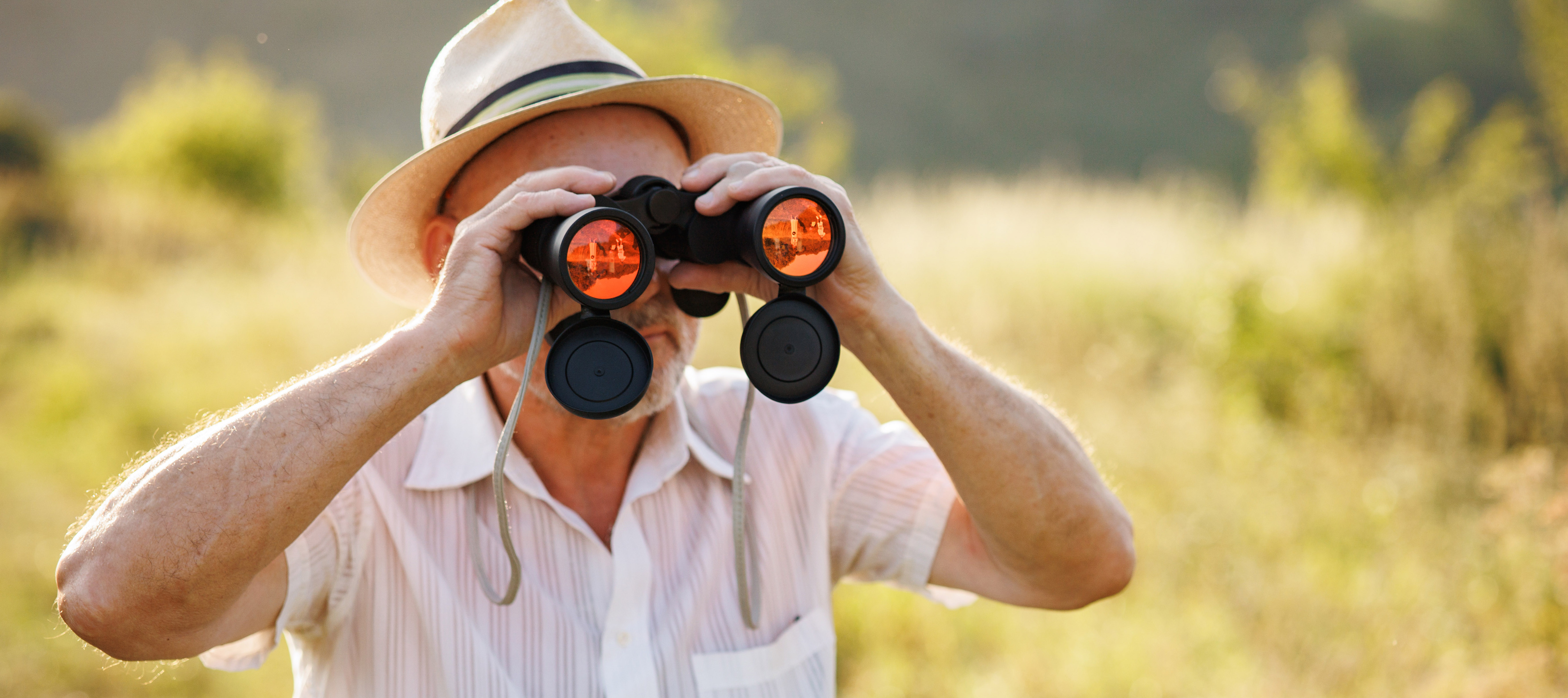 Portrait of a mature man with binoculars in a field. Bearded caucasian man looking through a binoculars in a field at summer. Man wearing striped t-shirt and a hat.