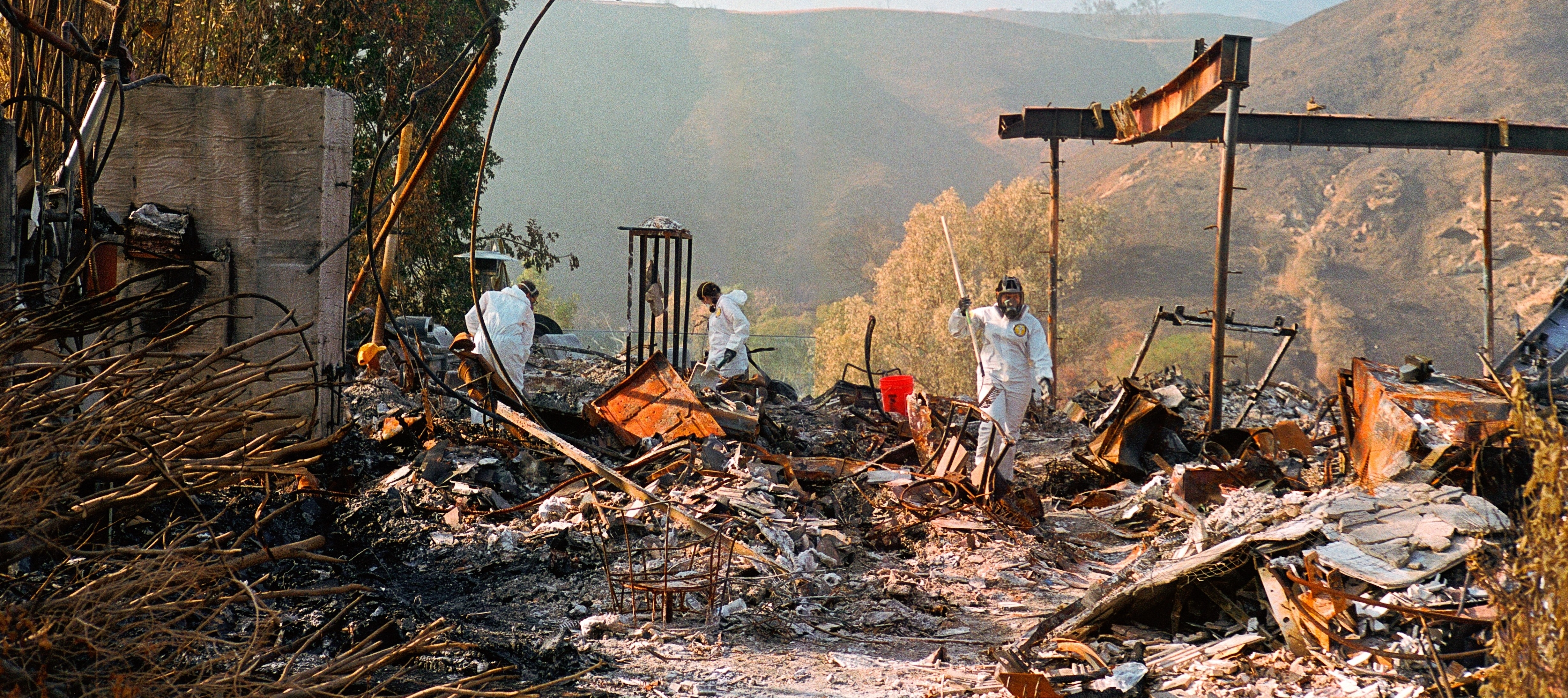 People in protective suits dig through the rubble of a home destroyed in the LA wildfires in 2025.
