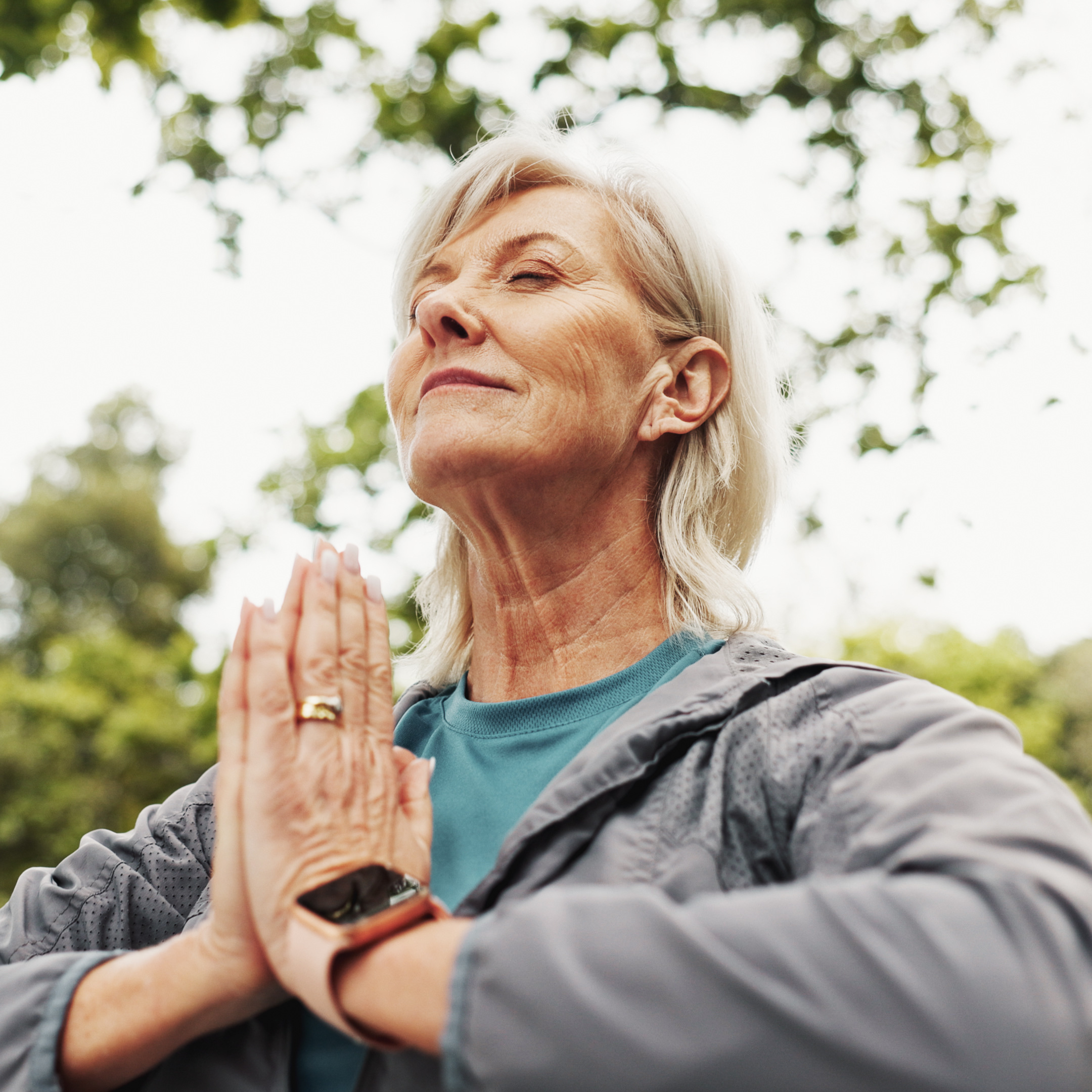Close up of an older woman outside with her hands pressed together and eyes closed, meditating or doing yoga.