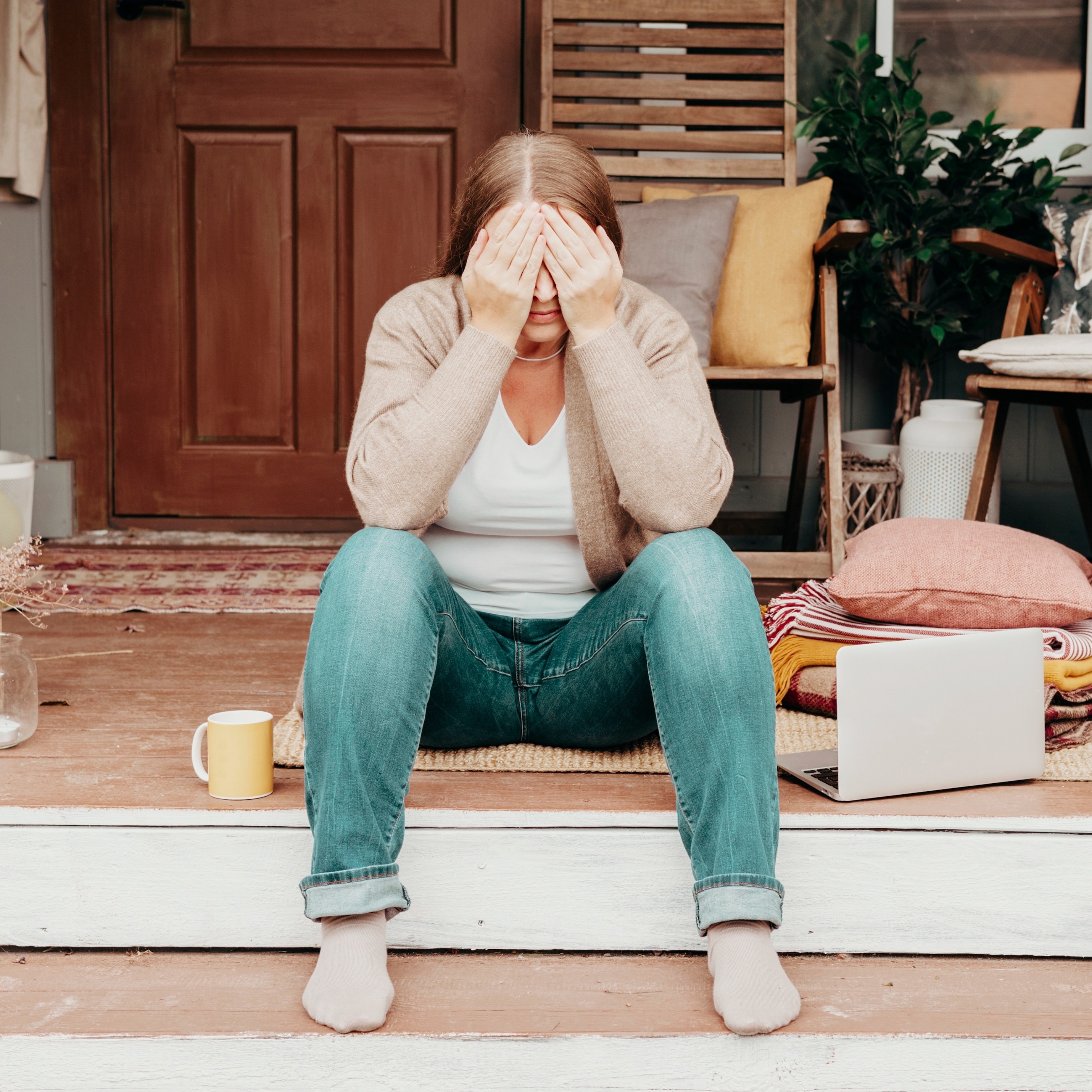 Woman sits on the front porch of her home with her hands over her face, looking stressed.