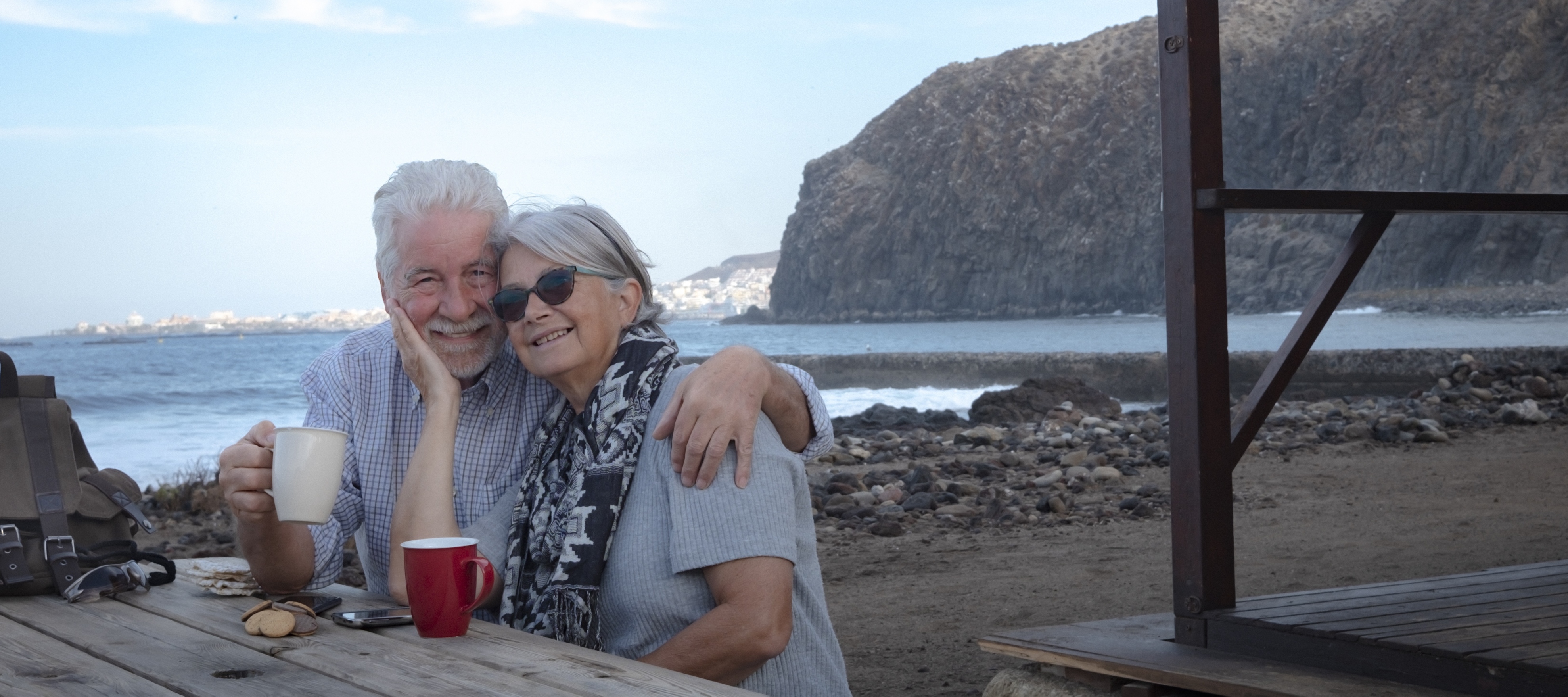 Retired couple enjoying coffee at a picnic table by the sea.