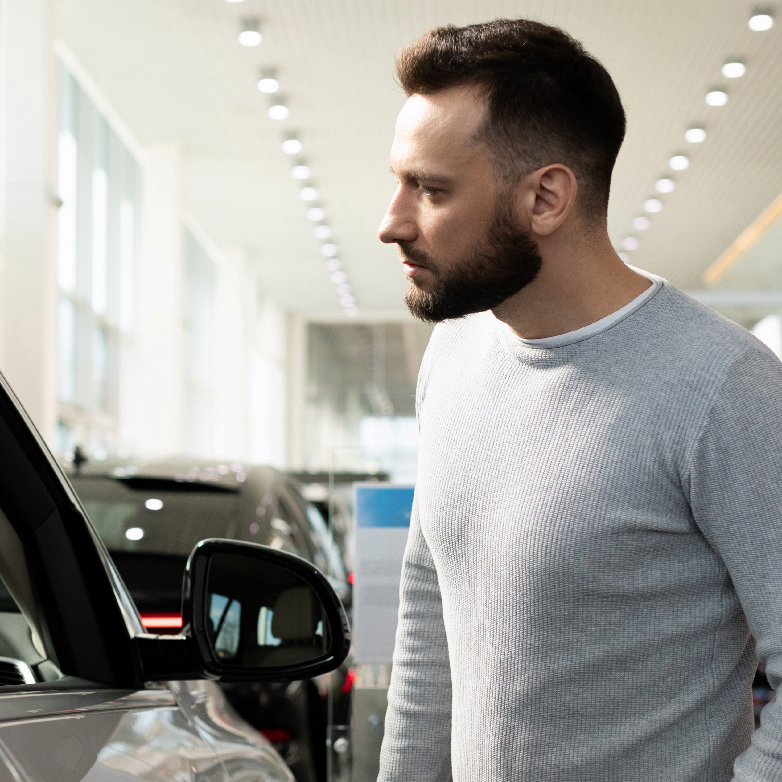 A man looks into the window of a new car while shopping at a car dealership.