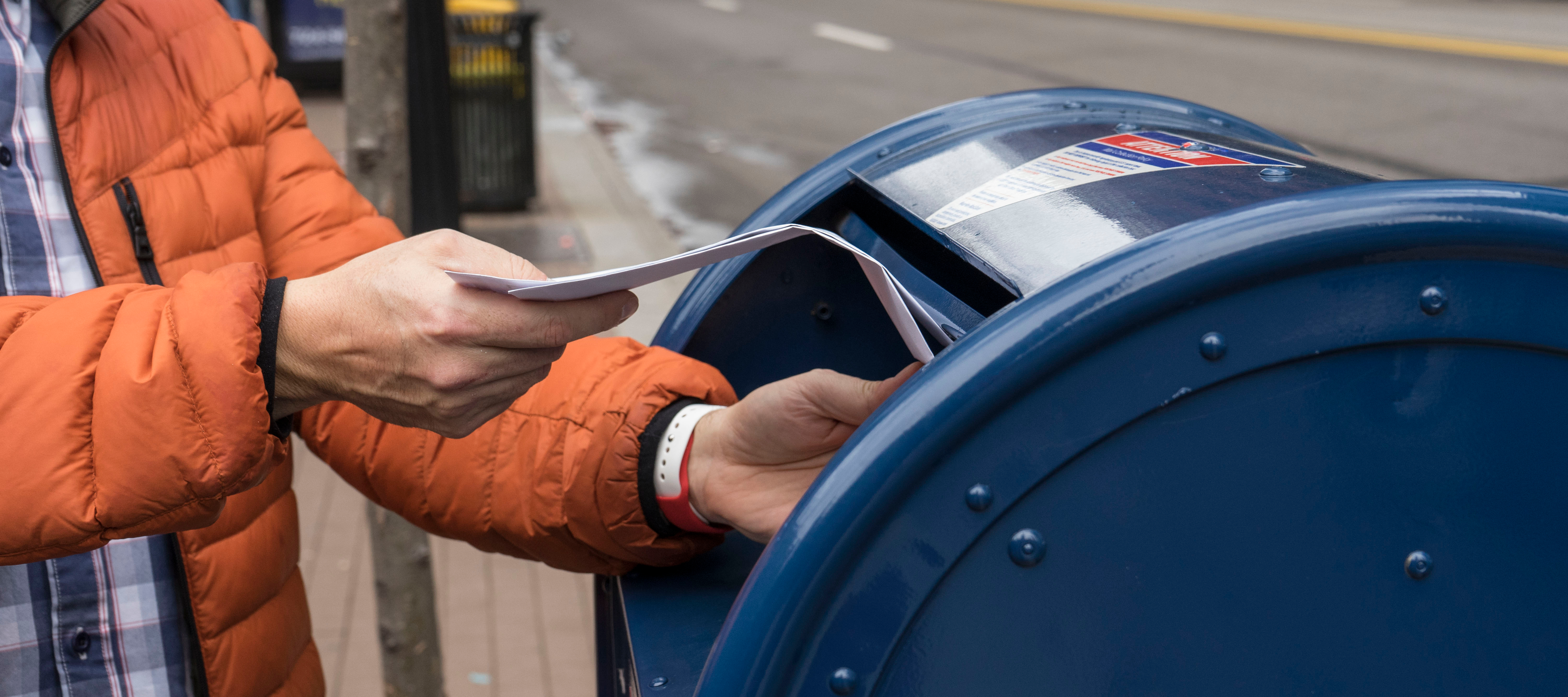 Close up of a person putting a piece of mail into the mail slot of a blue mailing box on a city street.