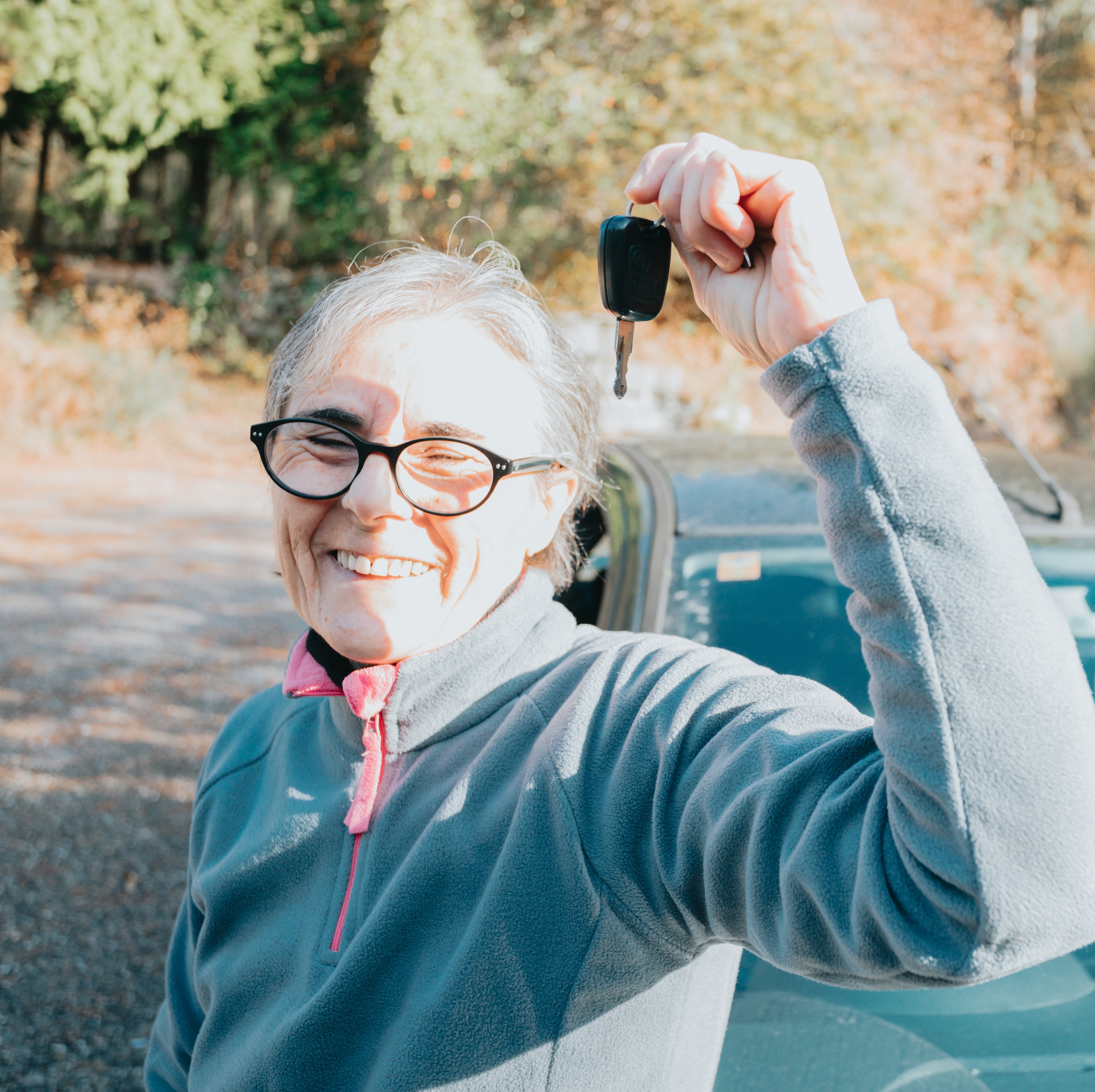 Older woman standing in front of new car in trail area, holding the keys and smiling.