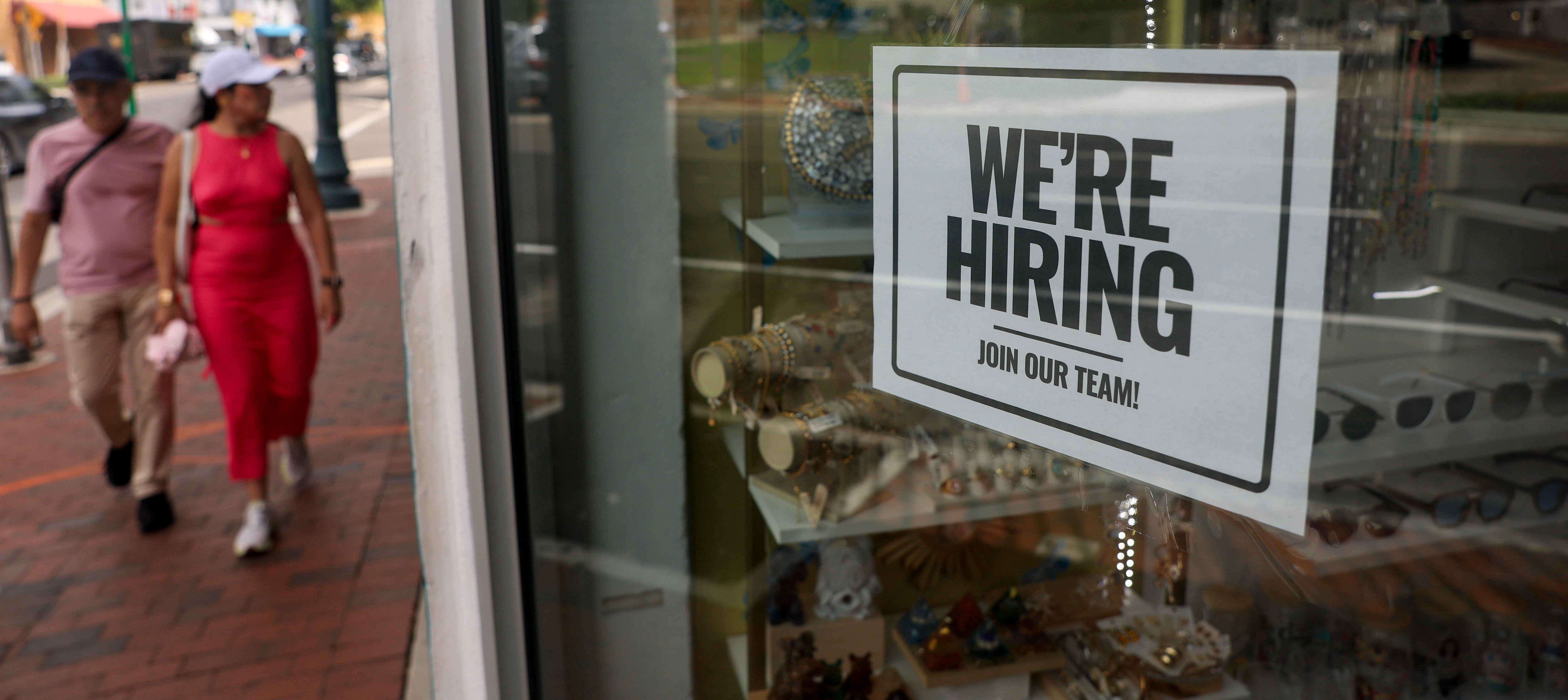 Two people walk by a store with a help wanted sign in the window.