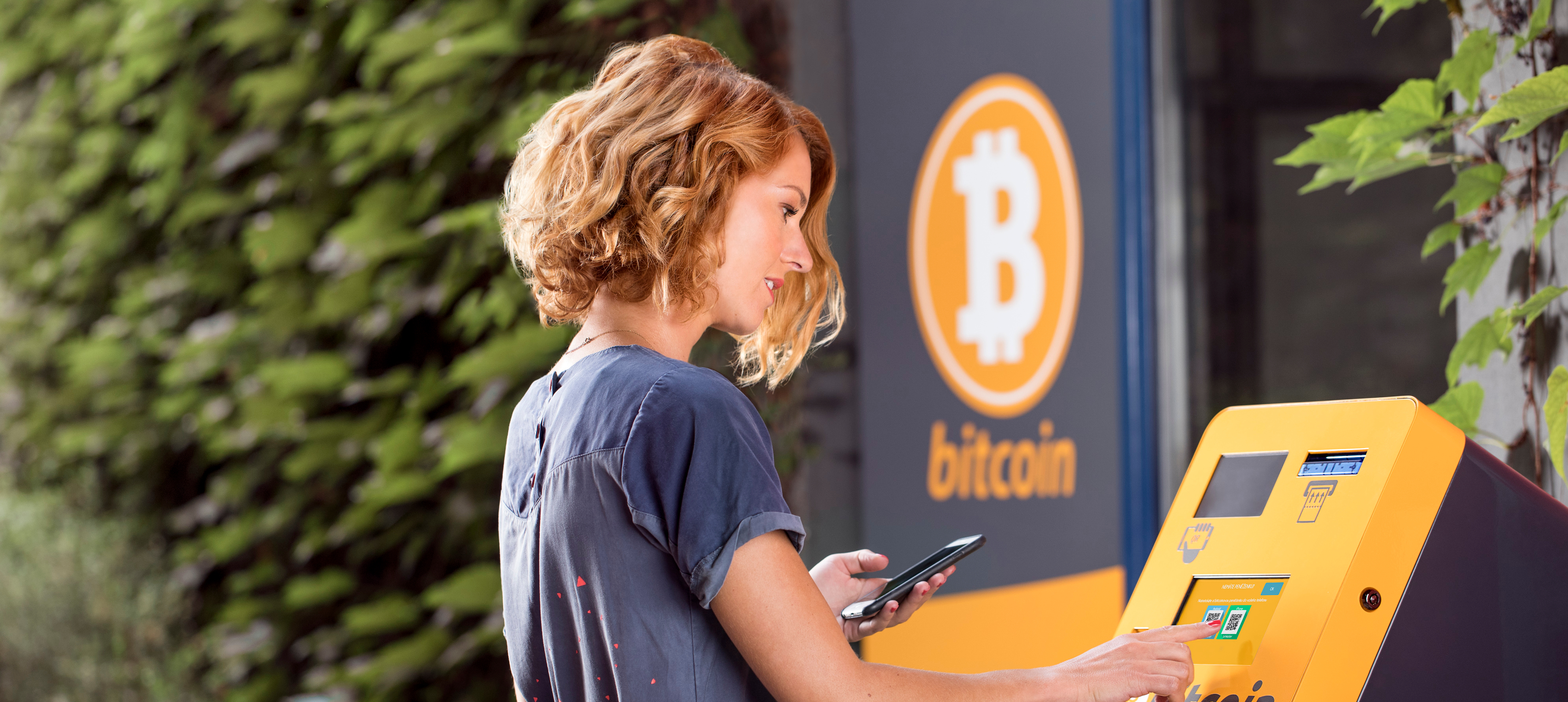 Woman in blue dress standing in front of yellow bitcoin ATM machine while holding phone, performing transaction.