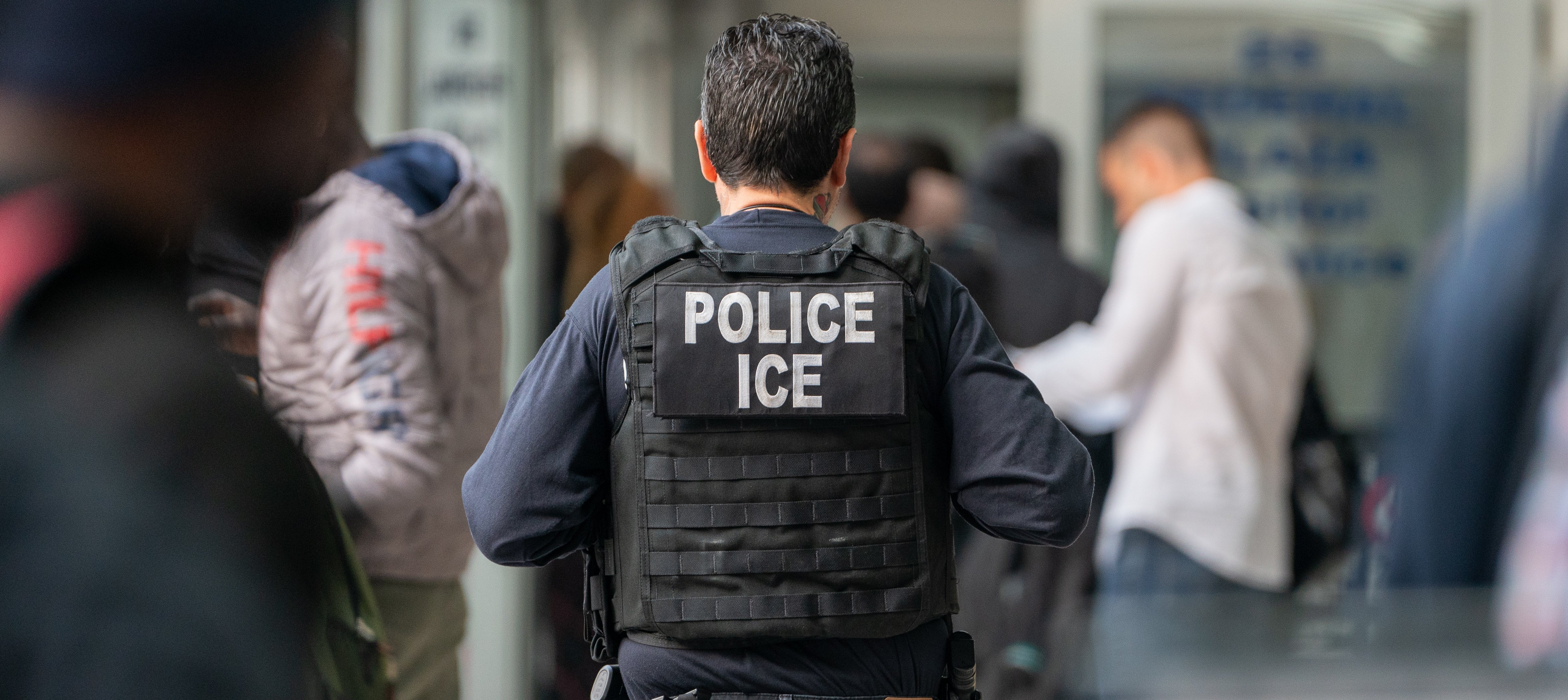 View from behind of an ICE agent monitors hundreds of asylum seekers being processed upon entering the Jacob K. Javits Federal Building
