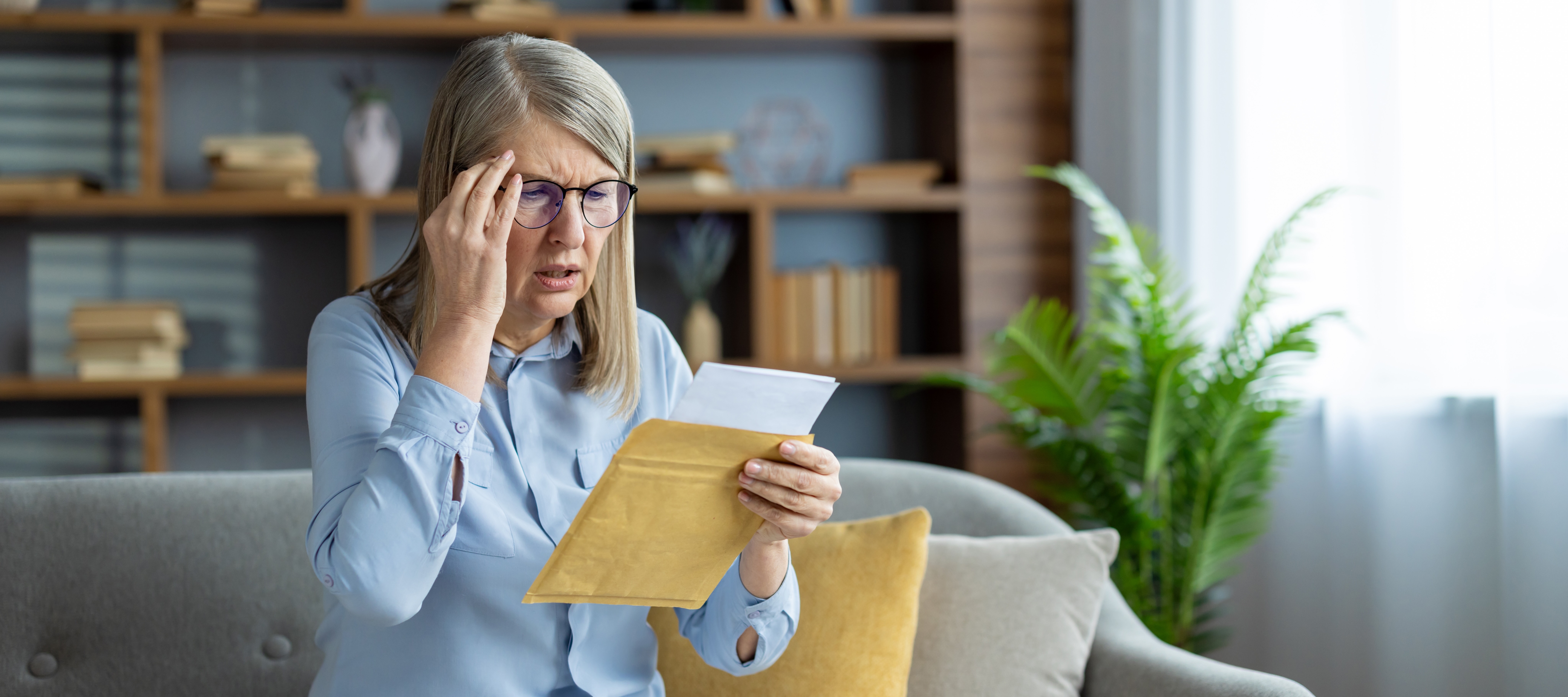 Woman sitting on couch looking confused and stressed while opening a piece of mail.