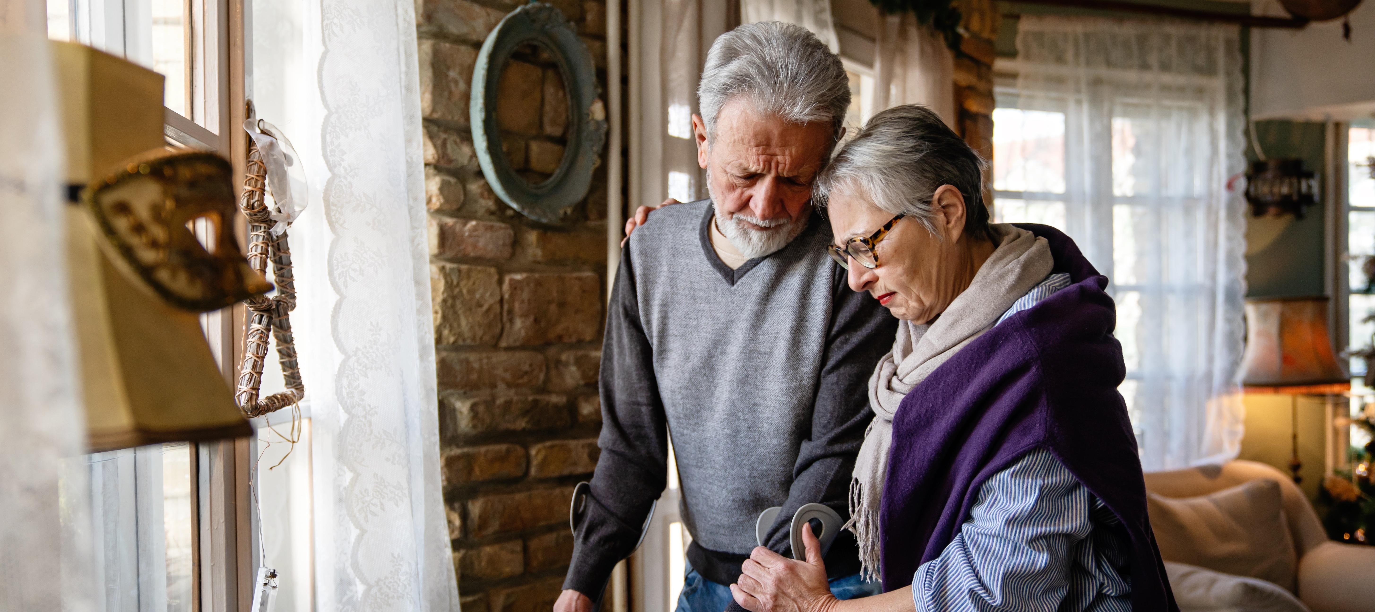 Elderly couple hold on to each other in home while one uses crutches to walk.