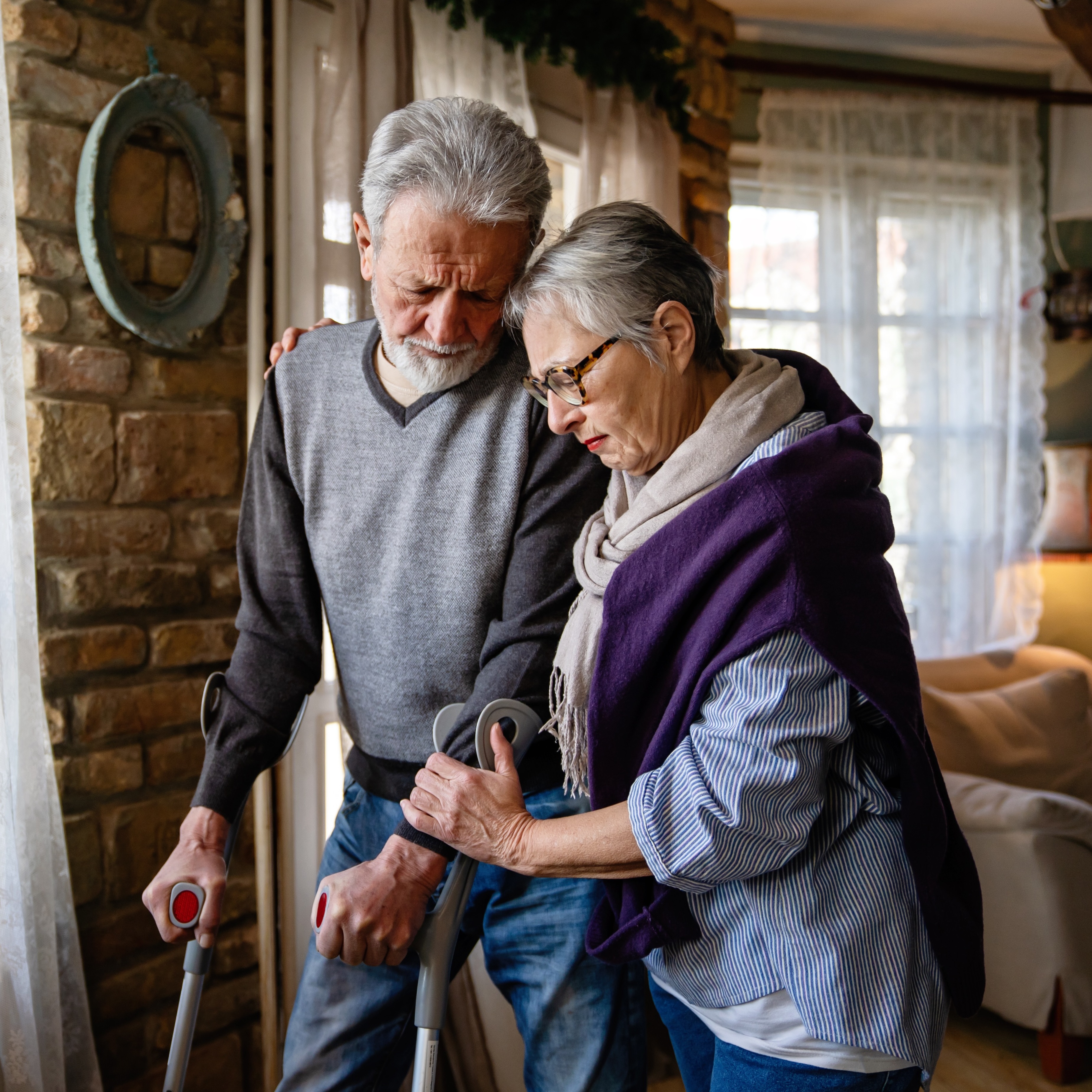 Elderly couple hold on to each other in home while one uses crutches to walk.