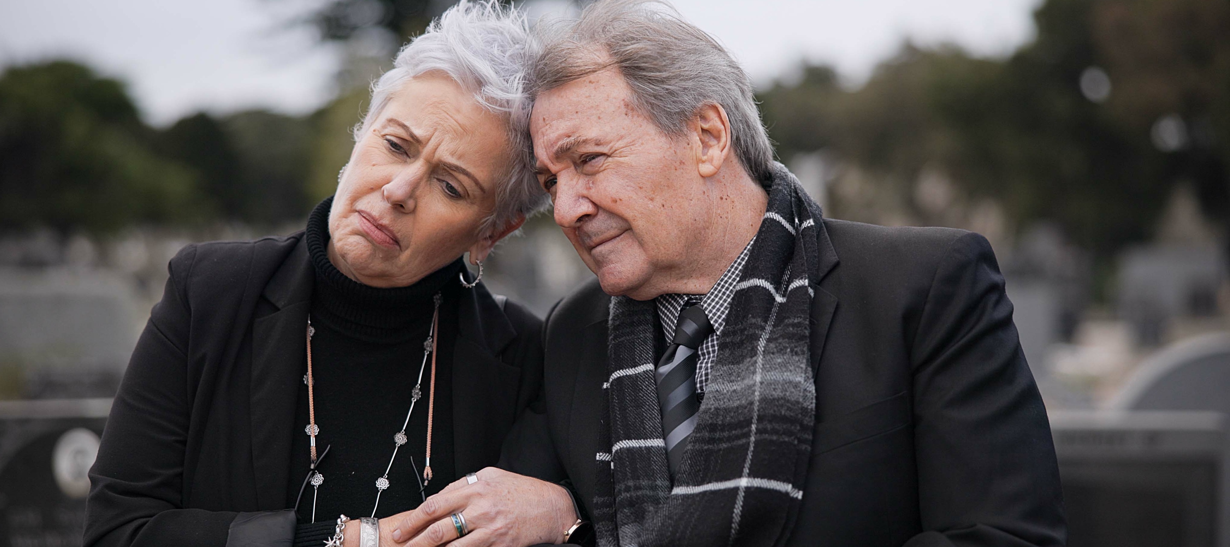 Older couple lean against each other in graveyard, looking distraught.