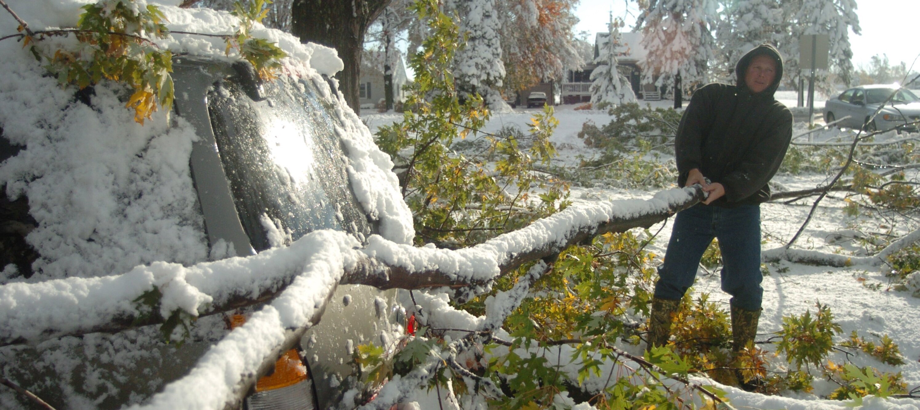 A man holds onto a large tree limb that has fallen on his car, covered in snow, and attempts to pull it off.