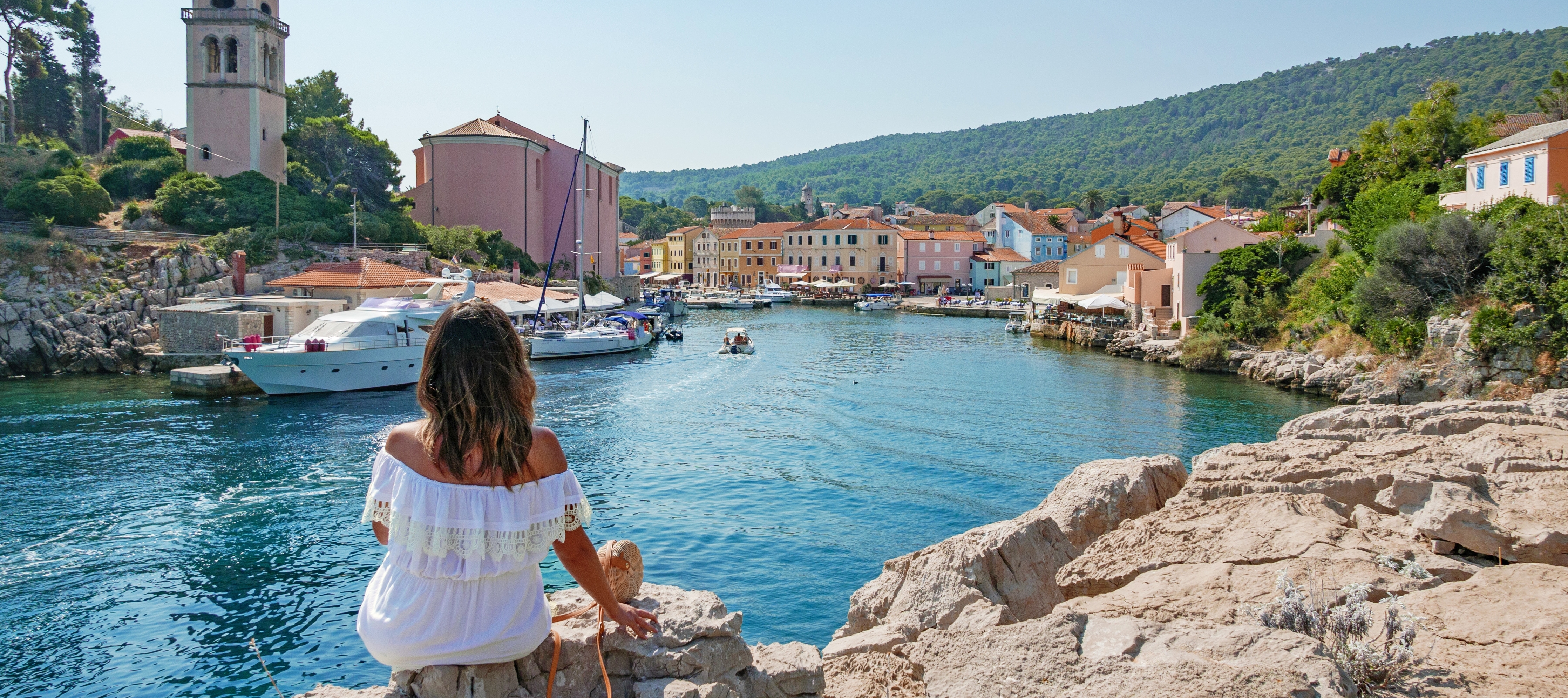 Rear view of young woman in white dress sitting on rock near an idyllic seaside town.