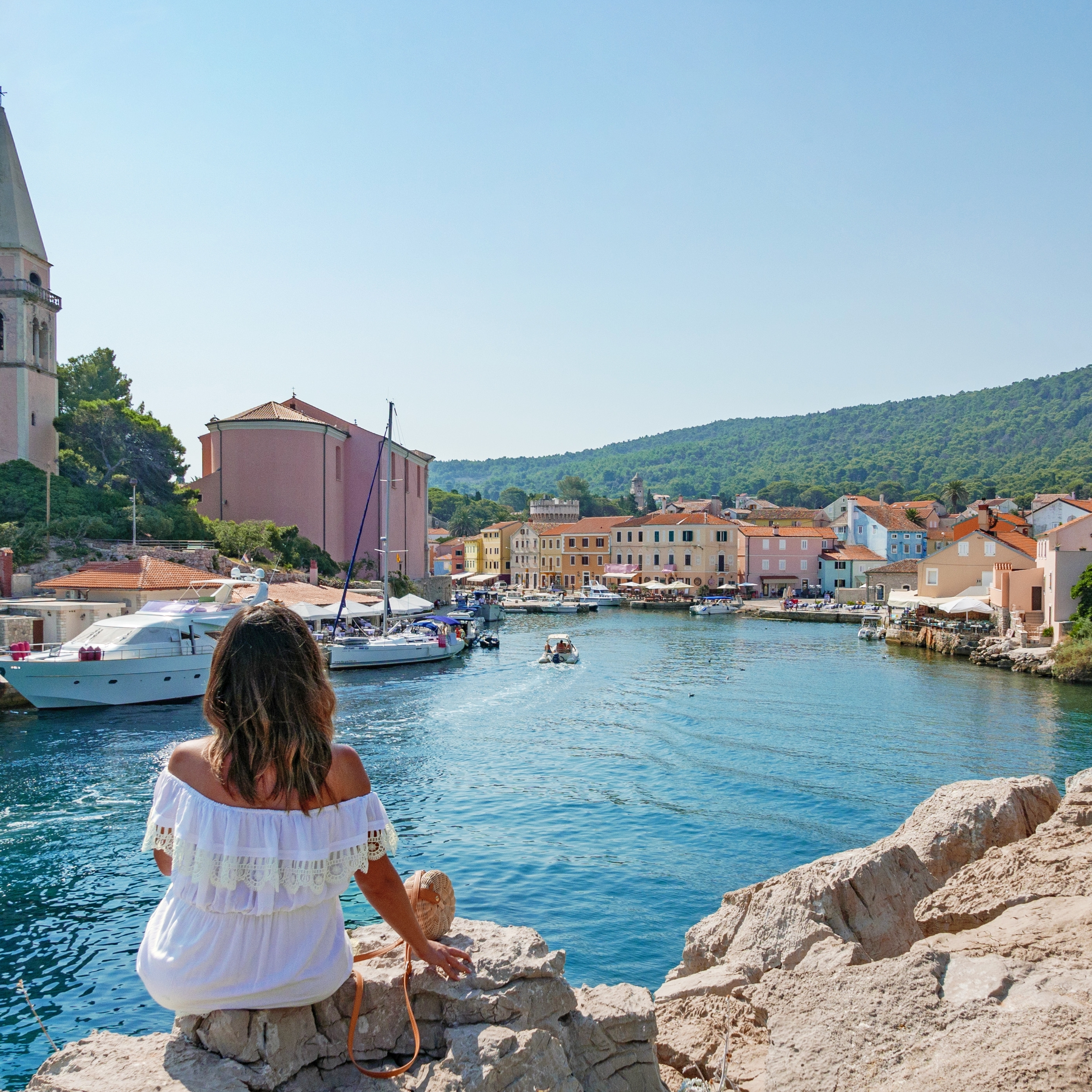 Rear view of young woman in white dress sitting on rock near an idyllic seaside town.