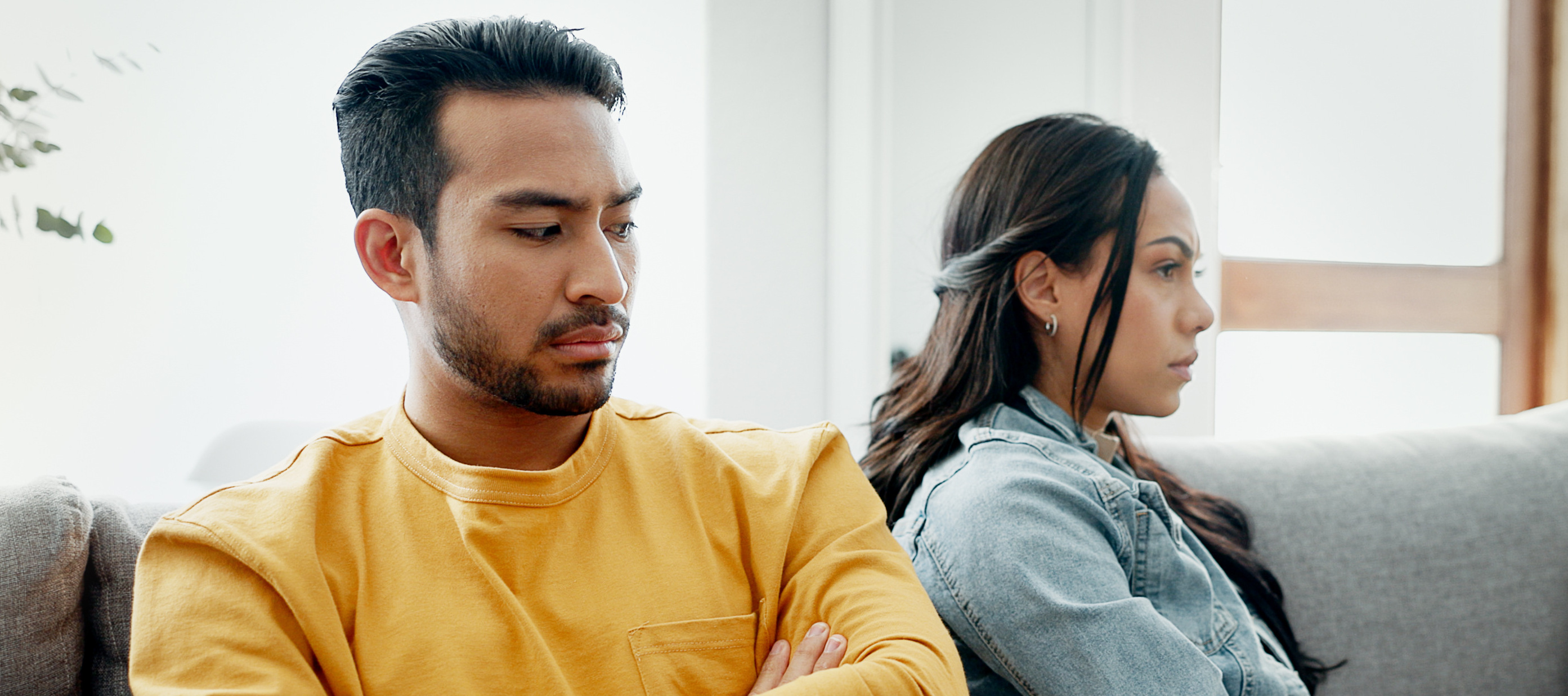 A couple sit on a couch mid-argument, looking away from each other.