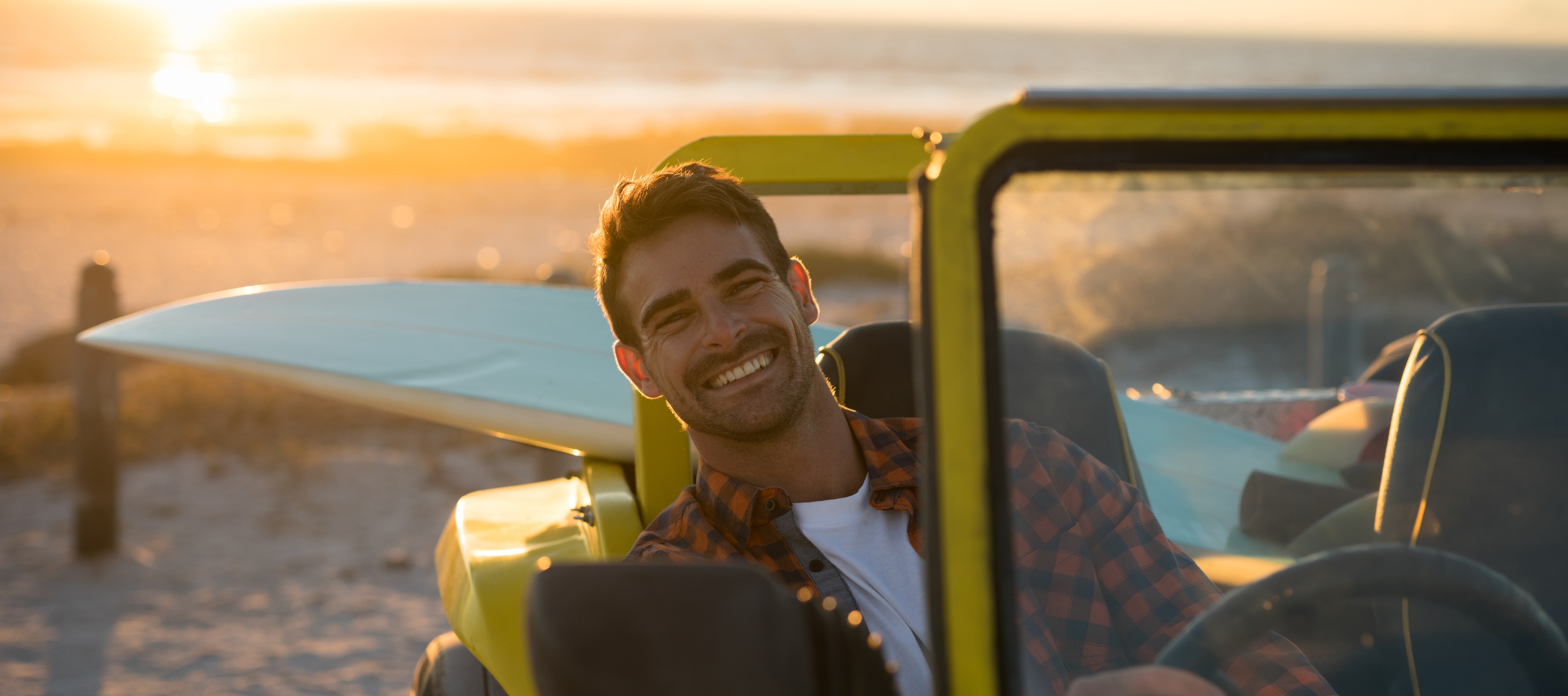 Smiling man sitting in car with open top on a beach with a surfboard in the back seat.