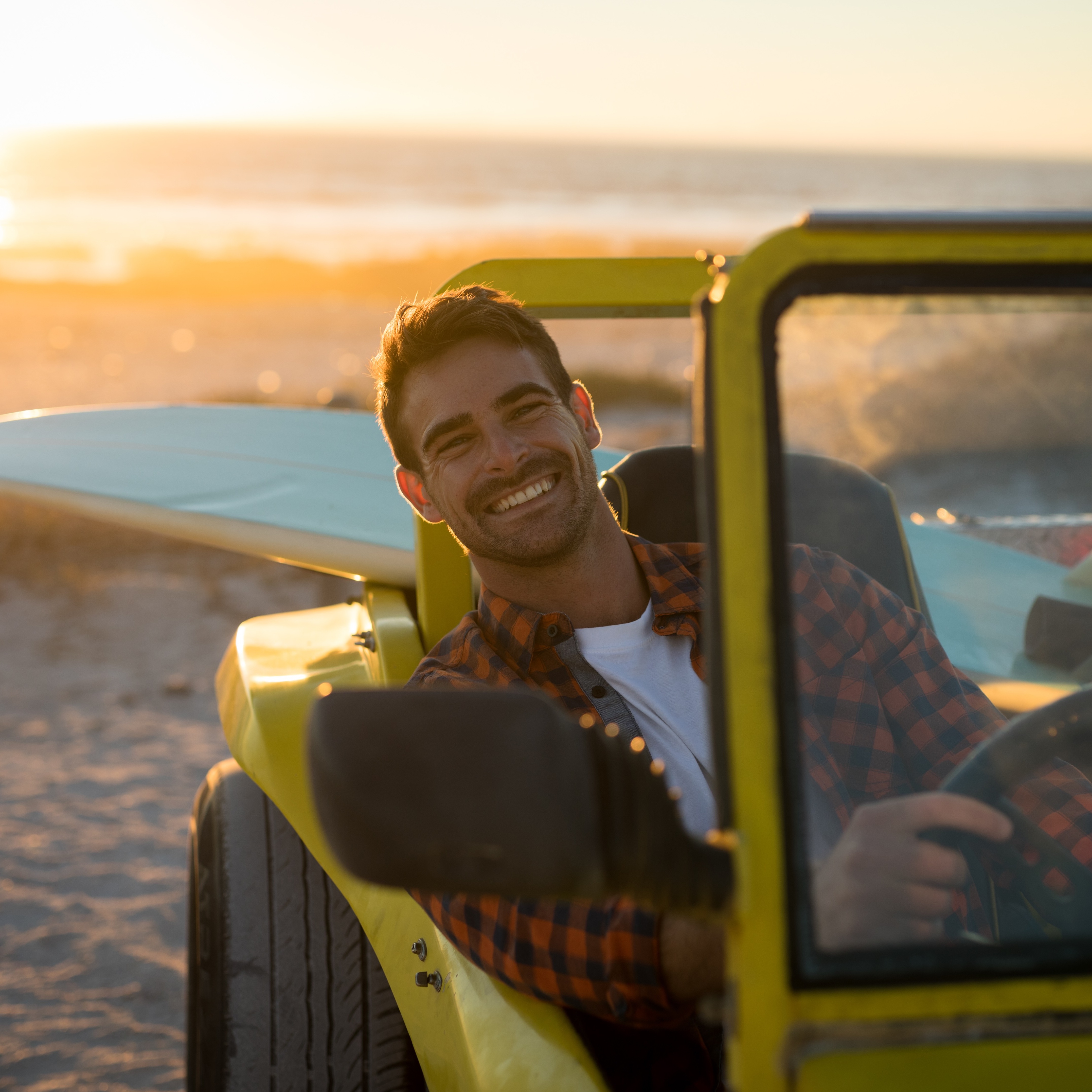 Smiling man sitting in car with open top on a beach with a surfboard in the back seat.