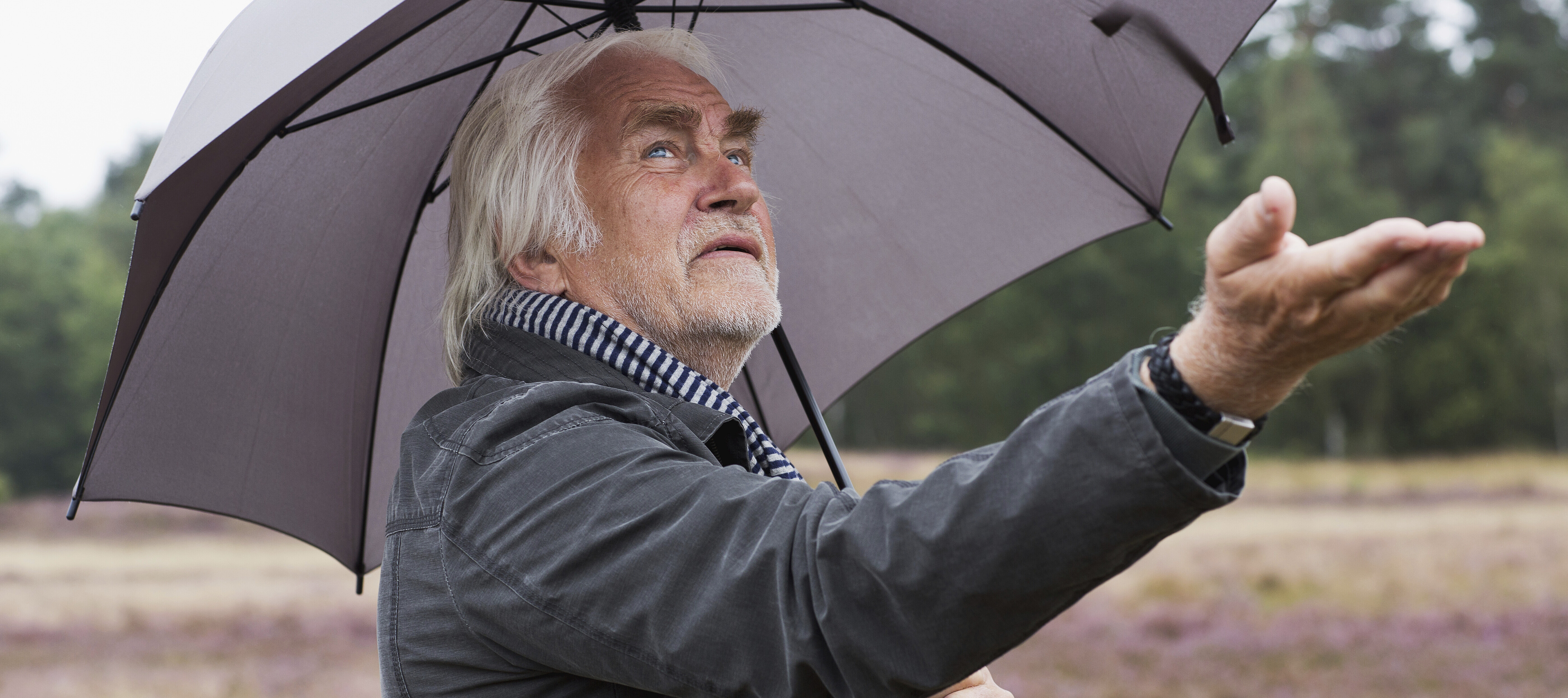 Senior man outside, standing under an umbrella, holding his hand out to feel for rain.