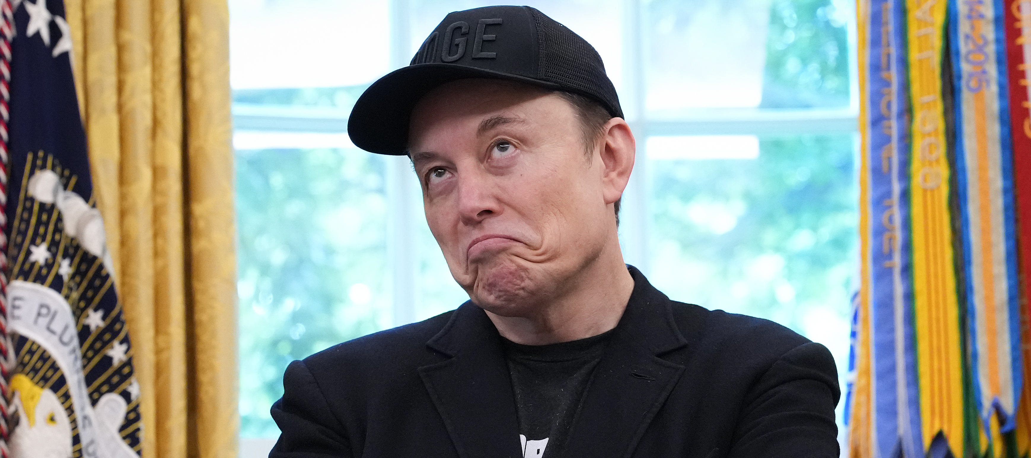 Elon Musk averts his eyes and grimaces, wearing a black T-shirt, suit jacket and DOGE baseball cap.