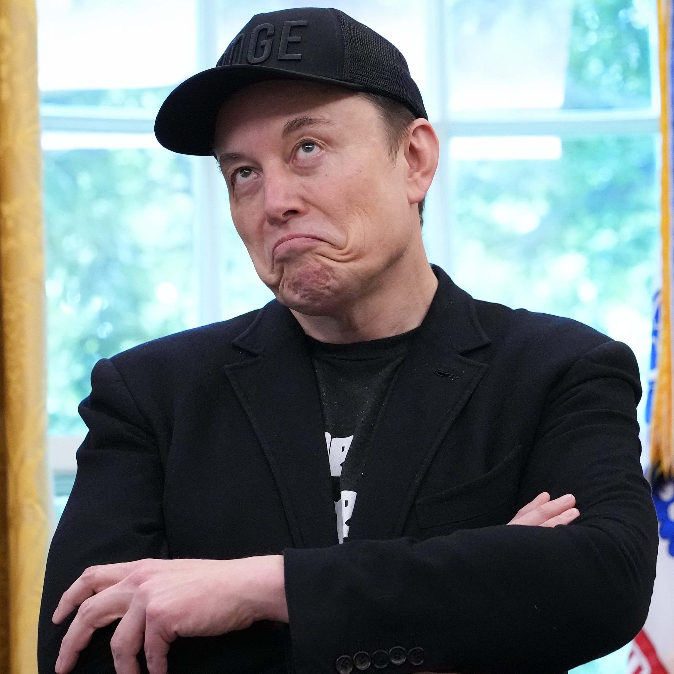 Elon Musk averts his eyes and grimaces, wearing a black T-shirt, suit jacket and DOGE baseball cap.