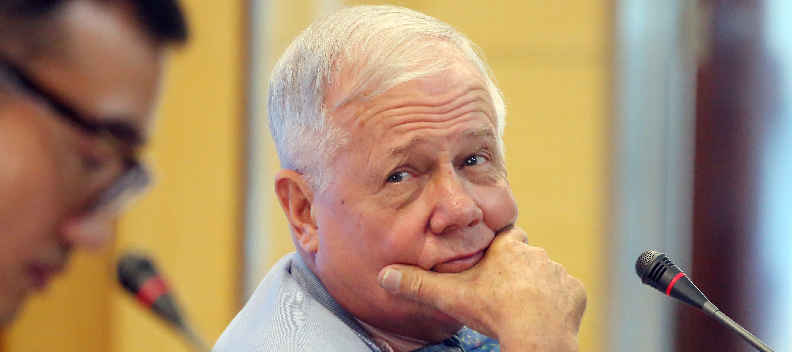 Jim Rogers sits at a table in a robin's egg blue suit, bracing the side of his jaw with one hand.
