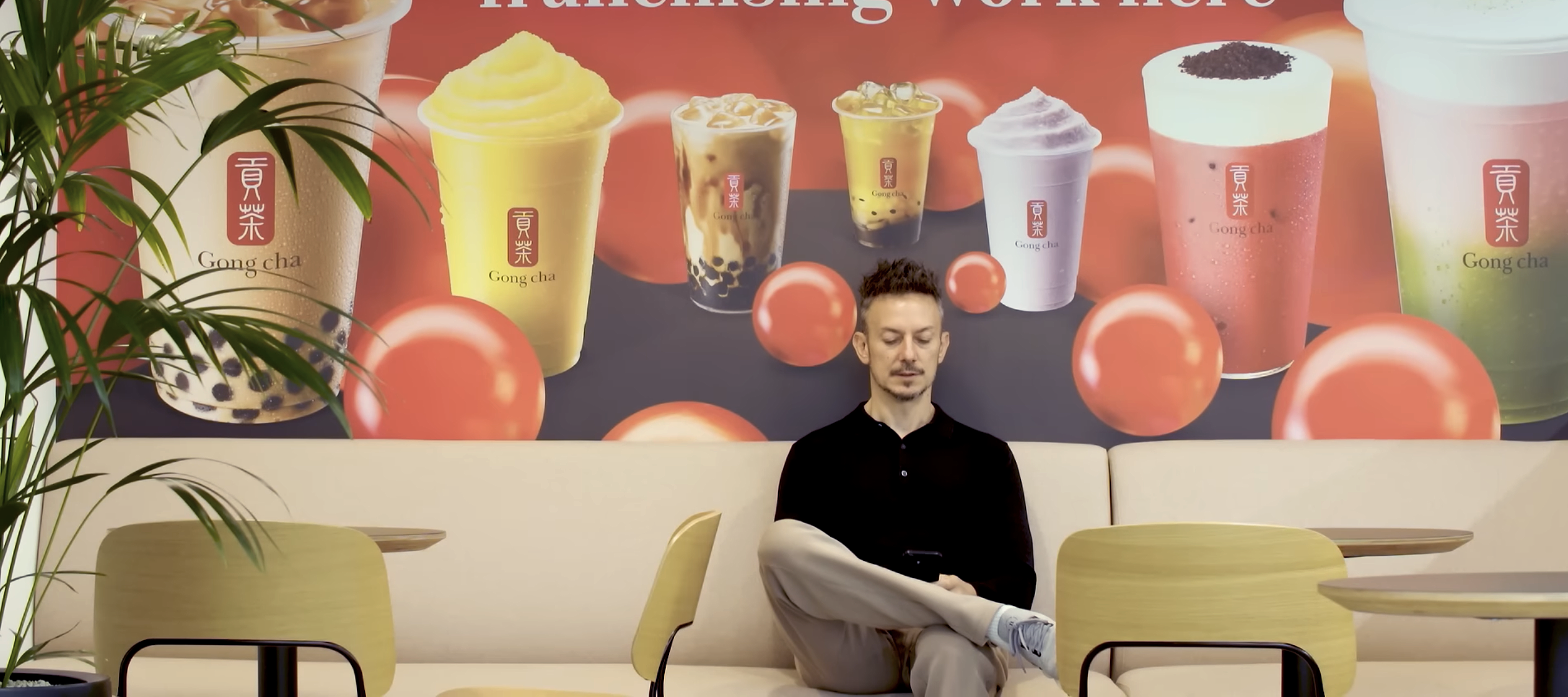 Martin Berry checks his phone while sitting in front of a Gong Cha poster.