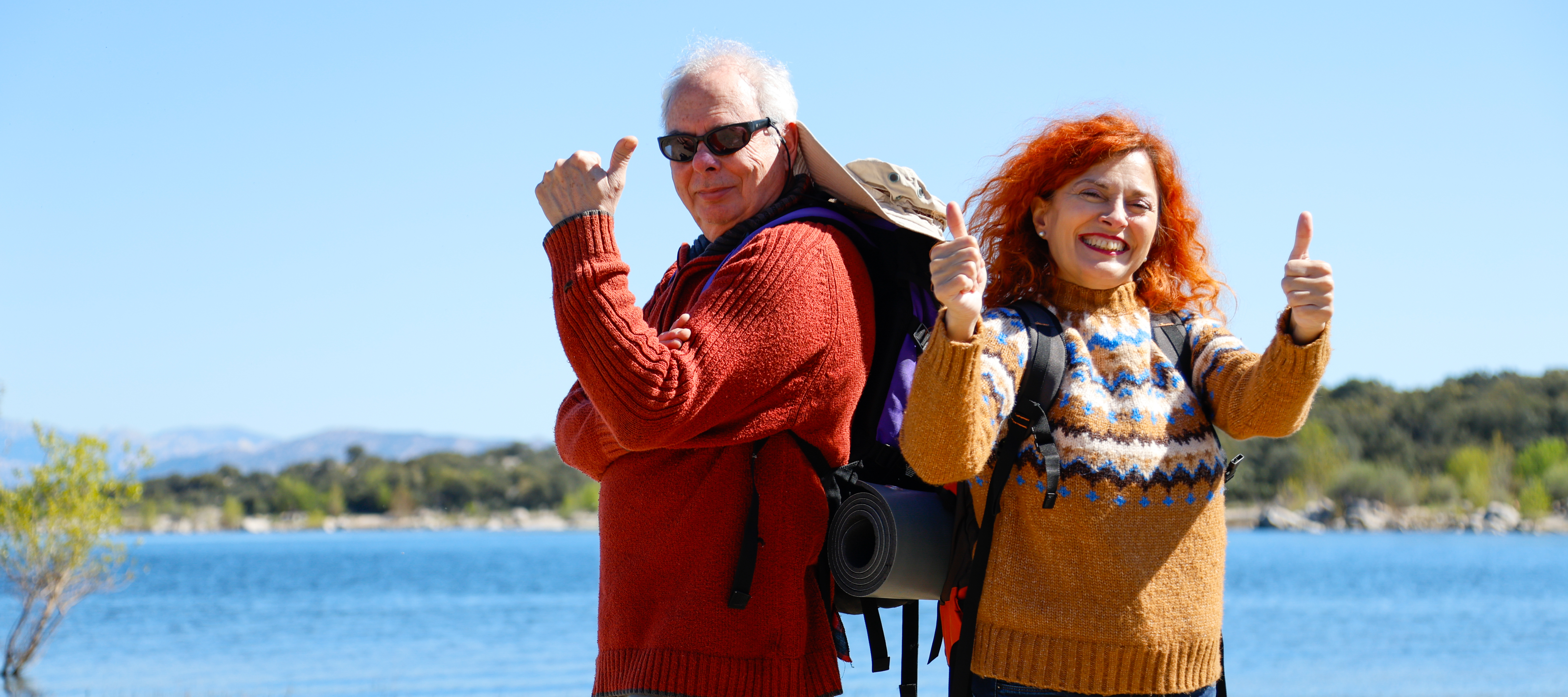 Happy senior couple with backpacks showing thumbs up while hiking near lake on a sunny day.