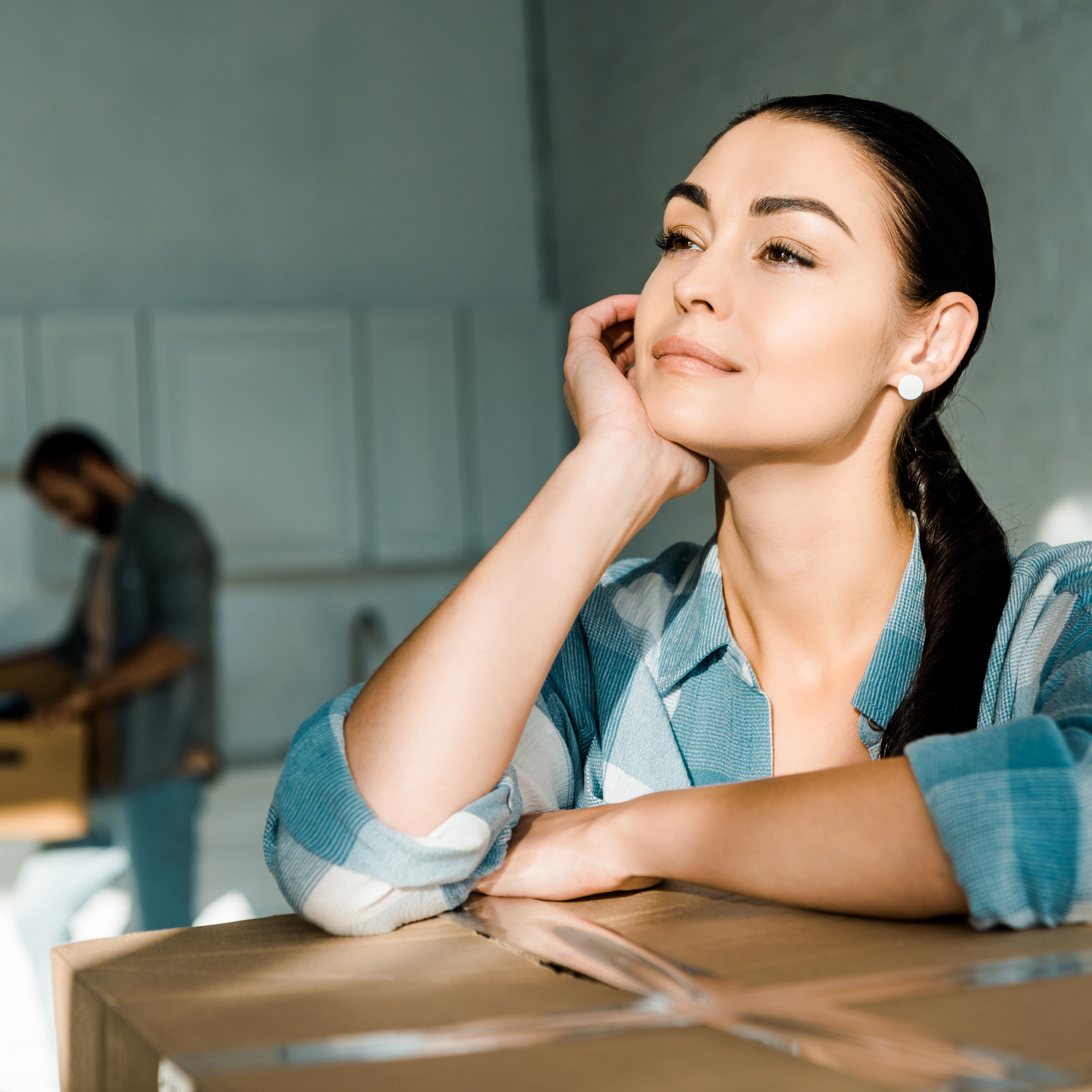 Woman leaning on top of moving box staring dreamily into the distance, while husband packs a box in the background.