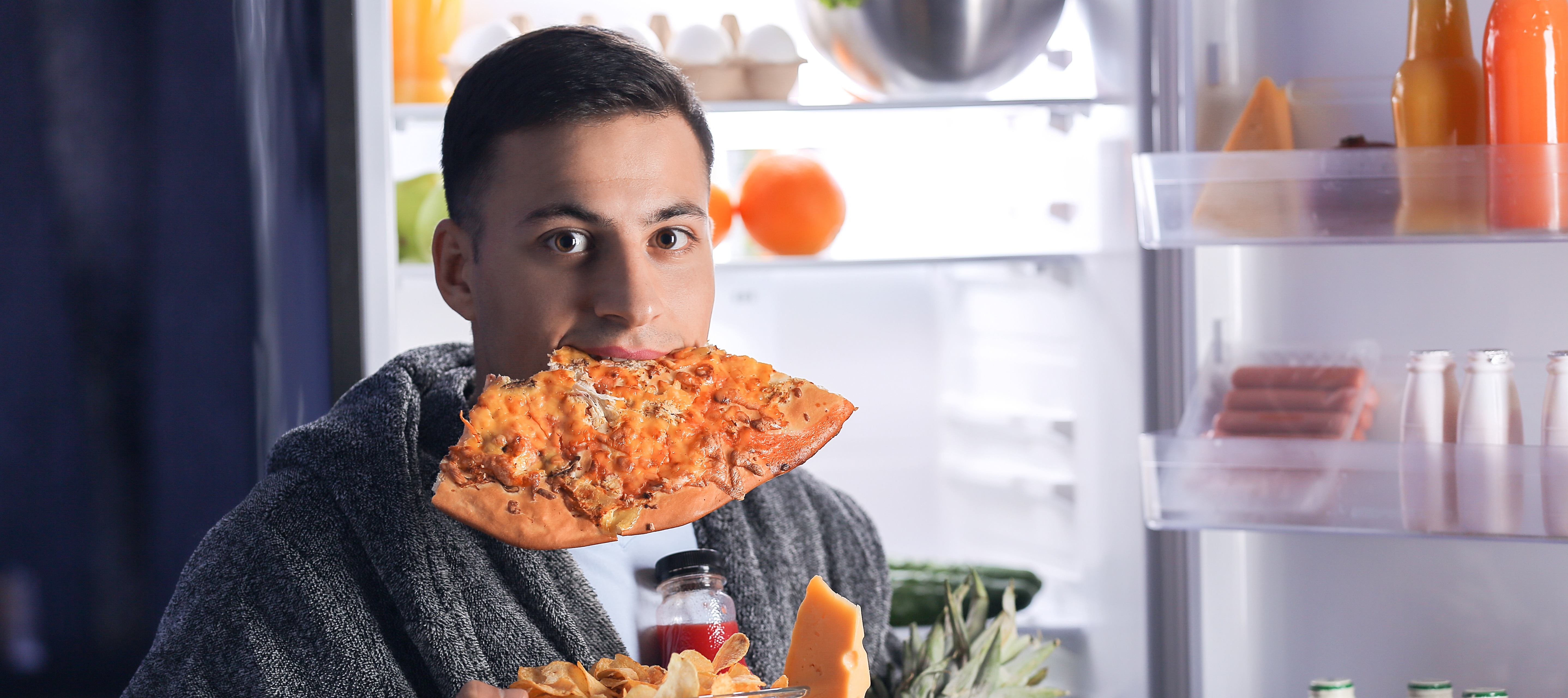 Adult with a piece of pizza in his mouth holding handful of other food standing in front of open refrigerator.