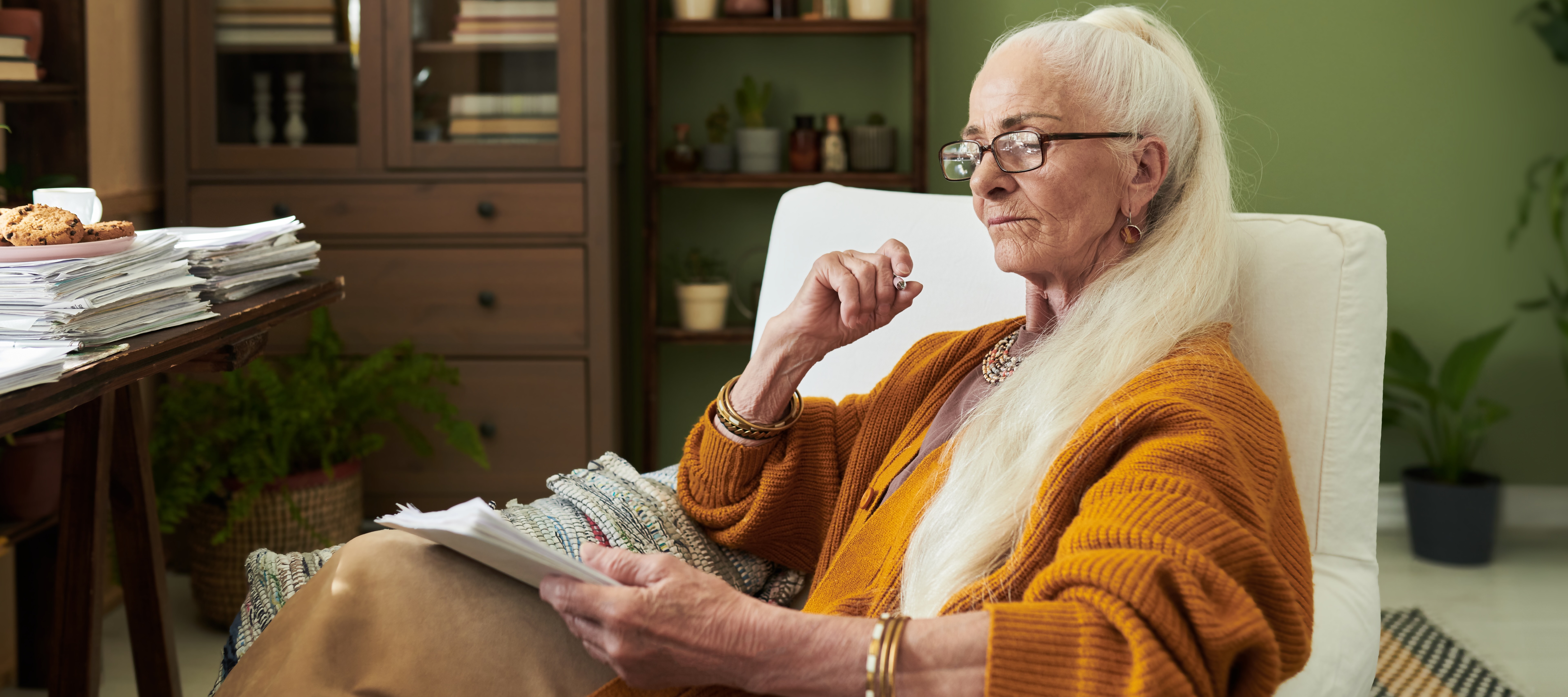 An older woman with long white hair sitting on a comfy chair in front of her desk and reviewing paperwork.