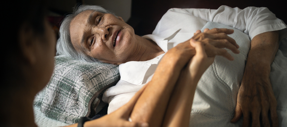 Daughter visiting her sick elderly mother in long-term care, lying in bed.