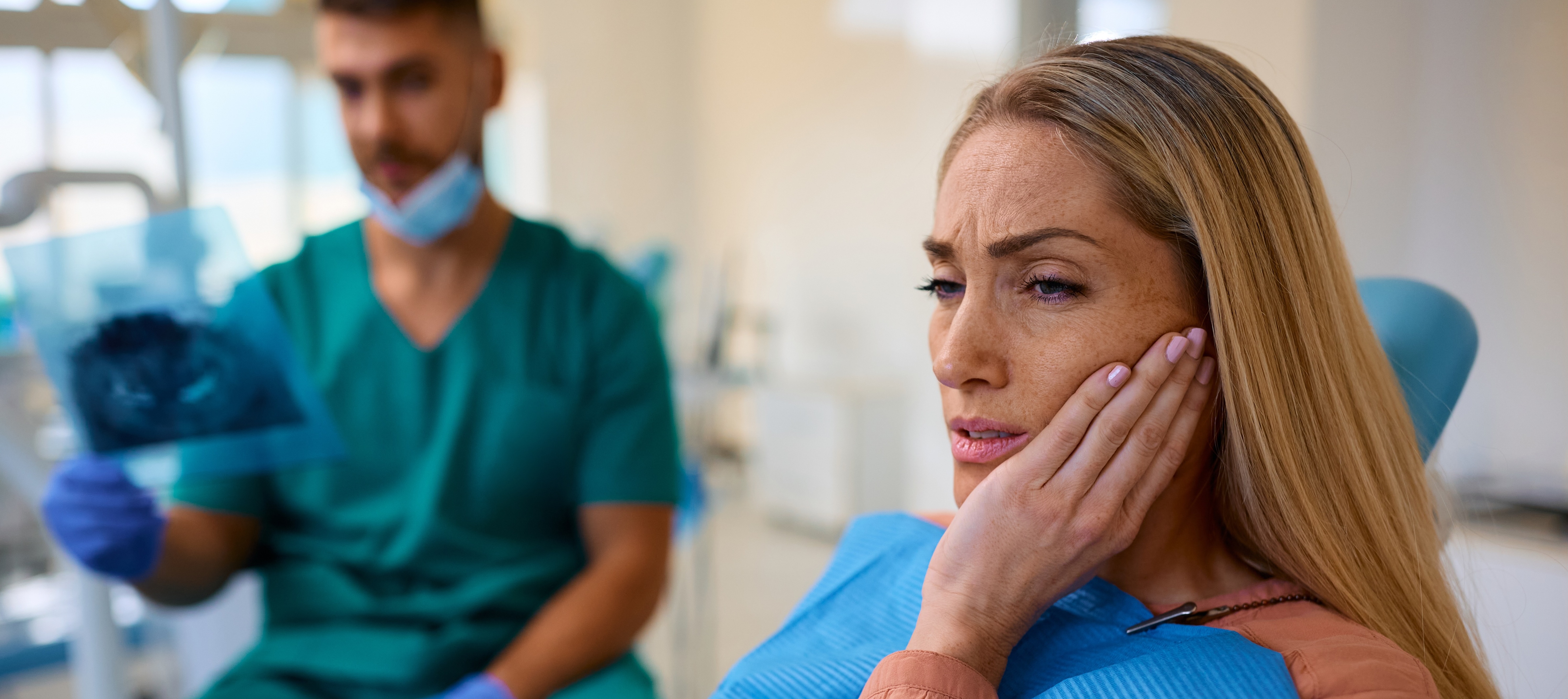 Distraught woman with toothache at dental clinic with her dentist in the background reviewing x-rays while she holds a hand to her face.