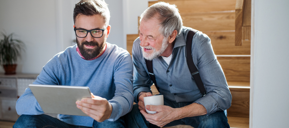 An adult millennial son and senior boomer father with tablet sitting on stairs indoors at home.