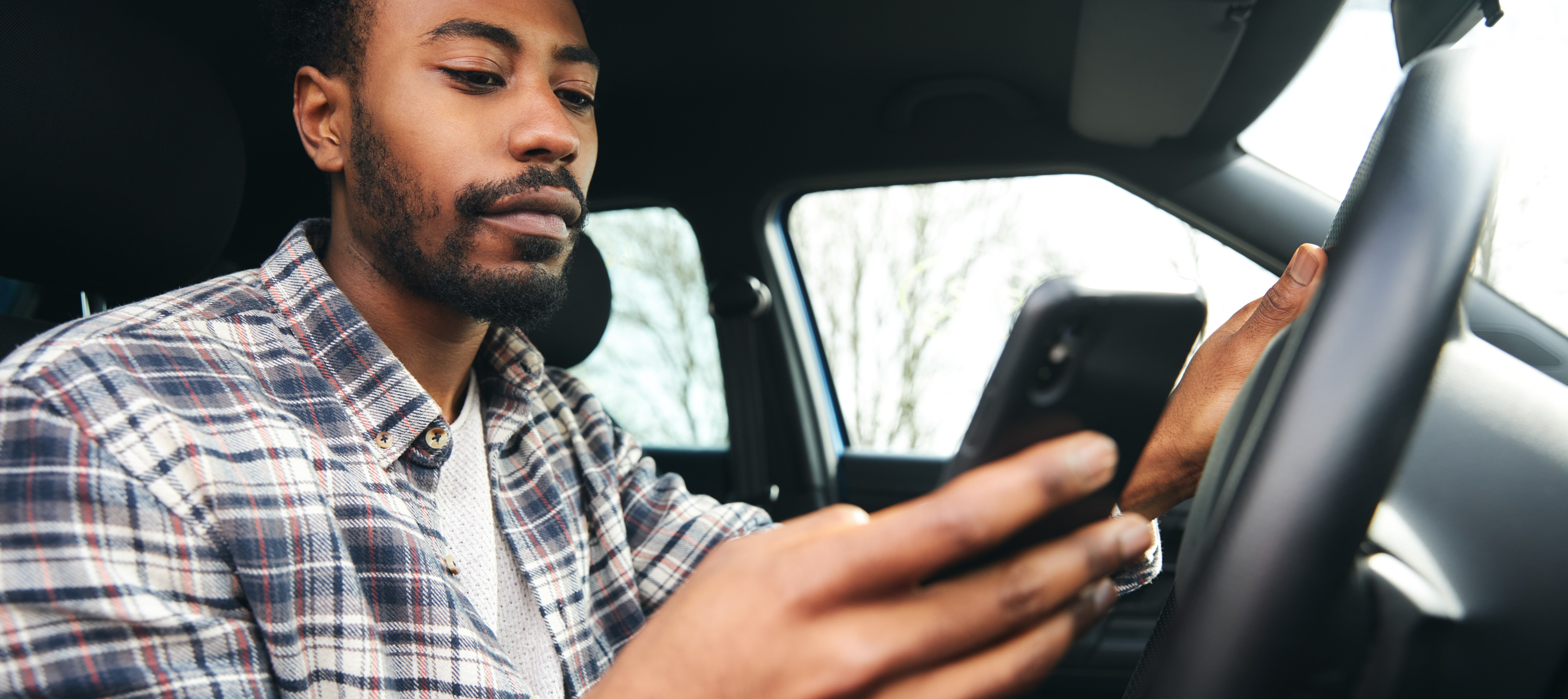 A man, sitting in a car, looks at his mobile phone, with AAA warning members about a scam offering "free roadside emergency kits."