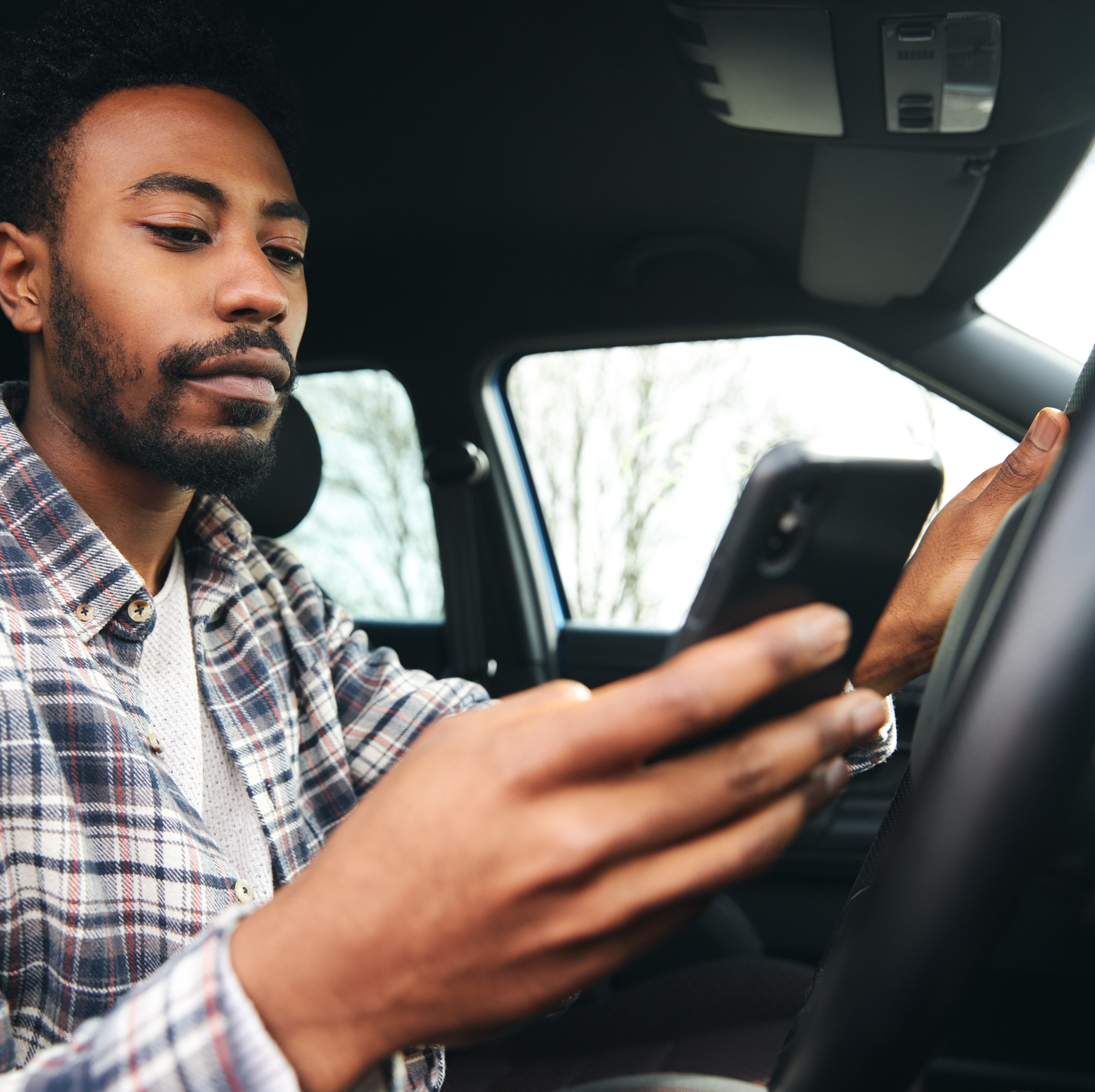 A man, sitting in a car, looks at his mobile phone, with AAA warning members about a scam offering "free roadside emergency kits."