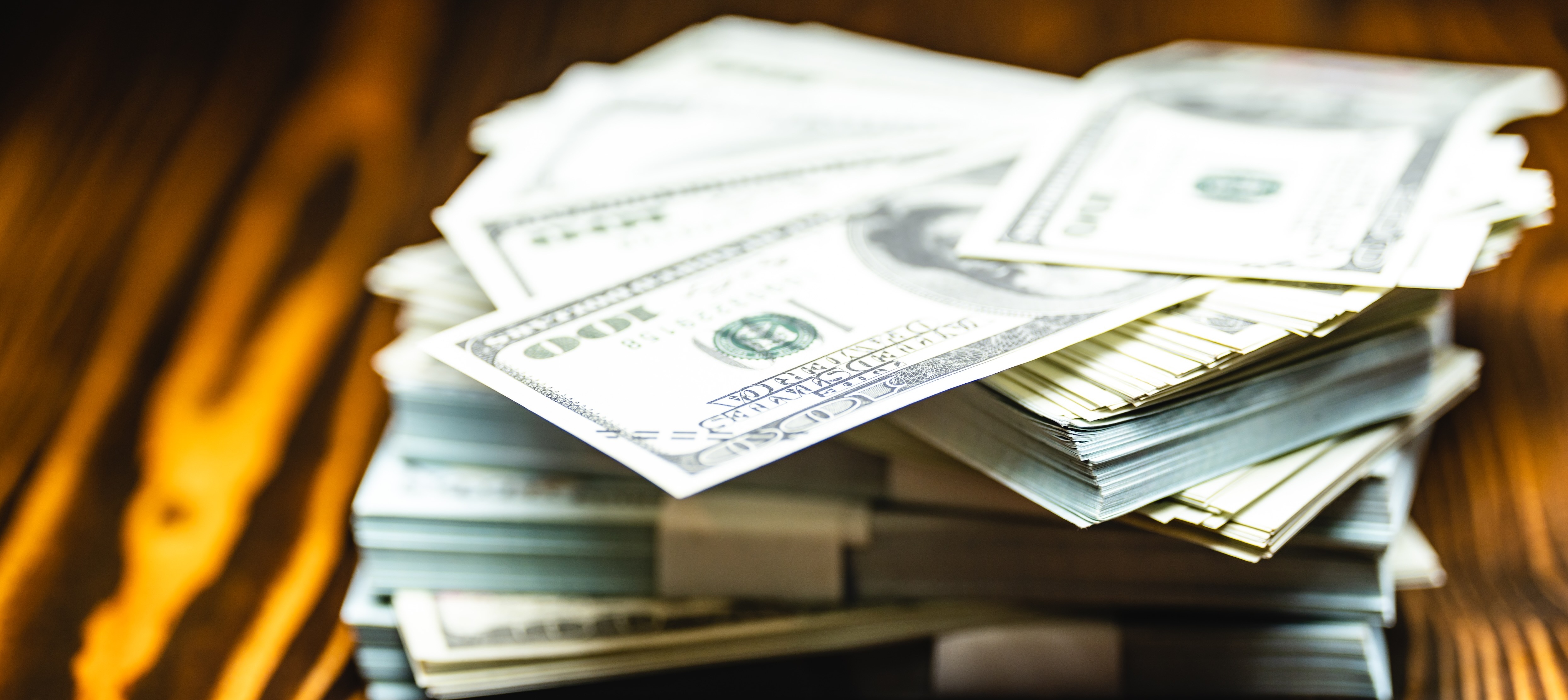 A close up shows multiple stacks of United States hundred dollar bills on a dark brown wood surface.