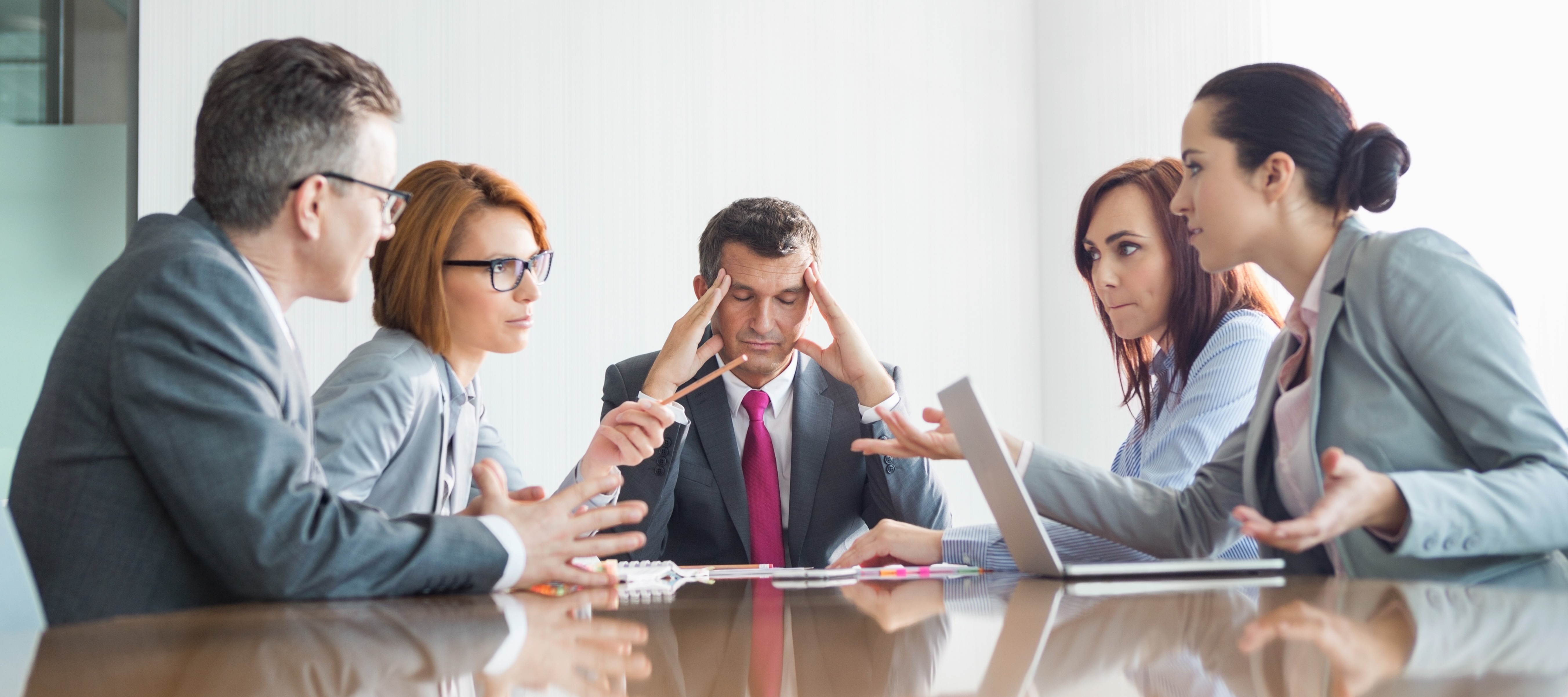 Businesspeople in a meeting having an argument while one man in the middle of it looks distressed, holding his head in his hands.
