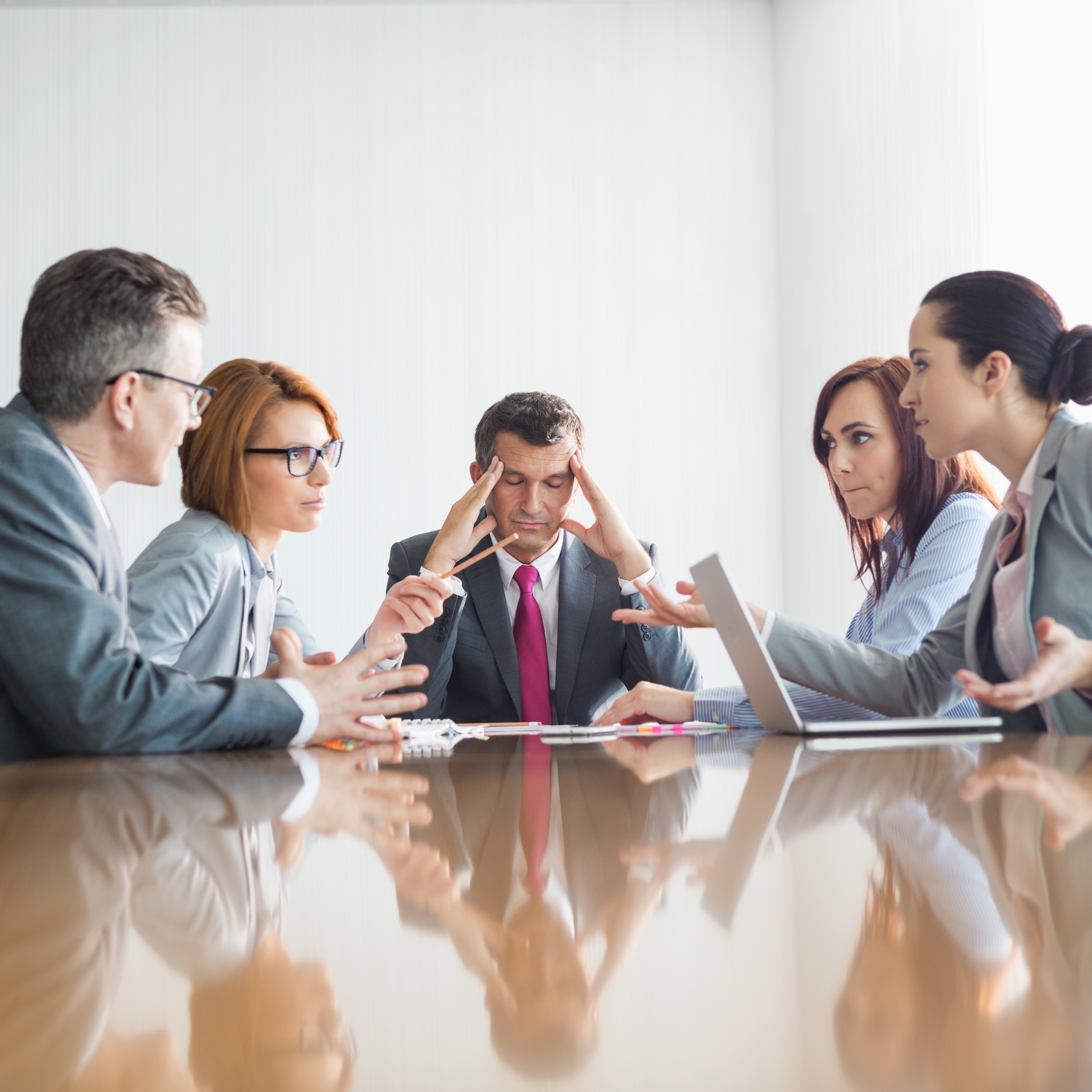 Businesspeople in a meeting having an argument while one man in the middle of it looks distressed, holding his head in his hands.