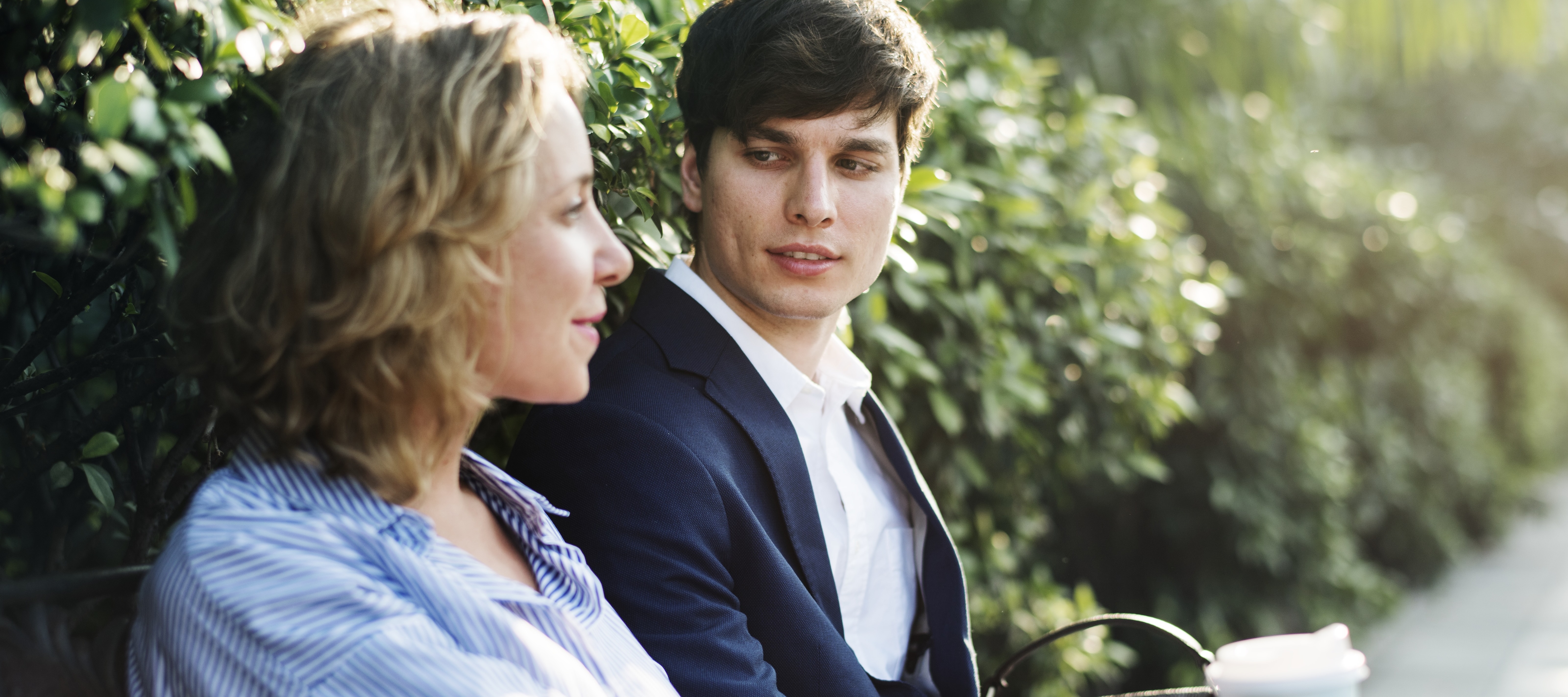 A mother and her college-aged son, sitting on a bench by a green bush, in deep conversation.