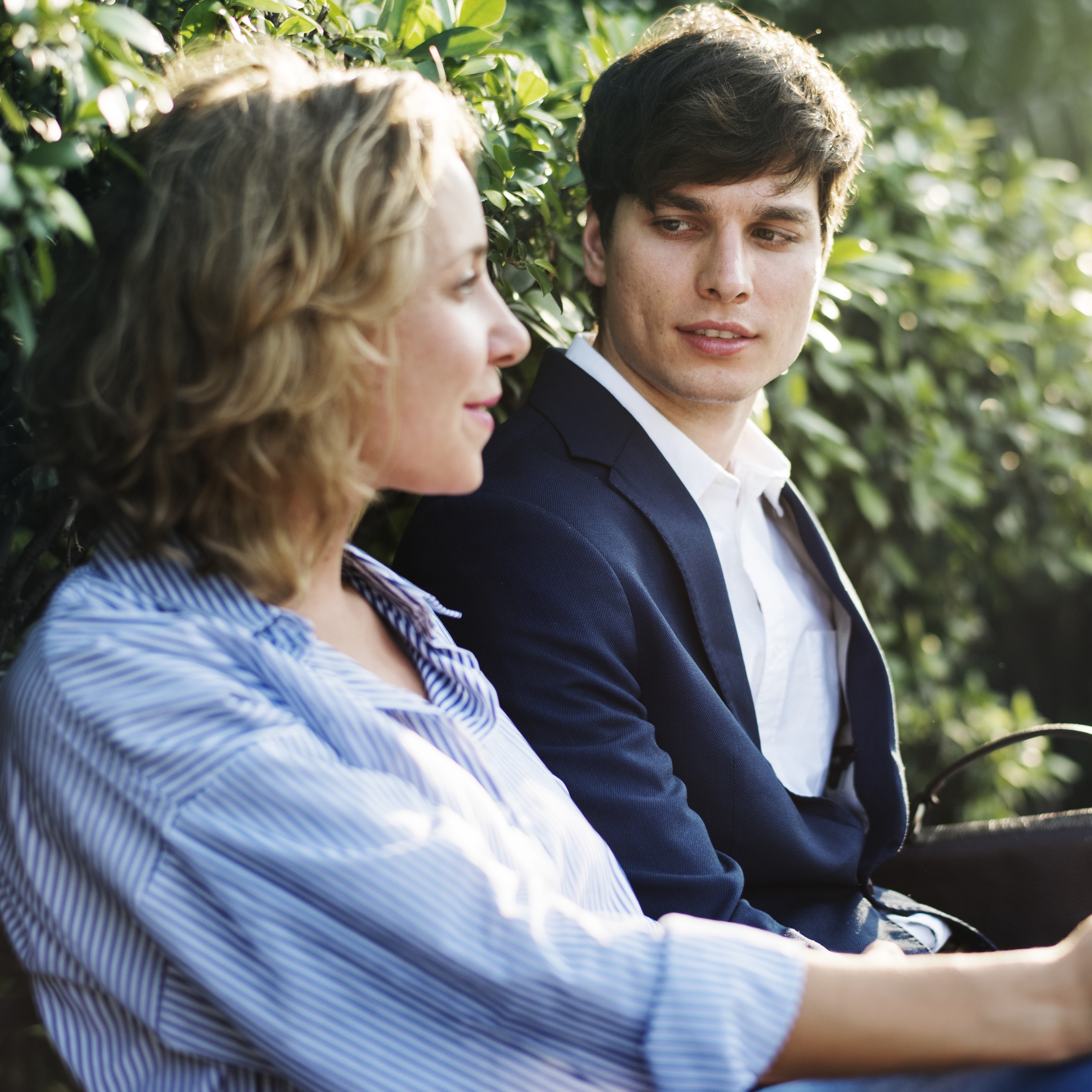 A mother and her college-aged son, sitting on a bench by a green bush, in deep conversation.
