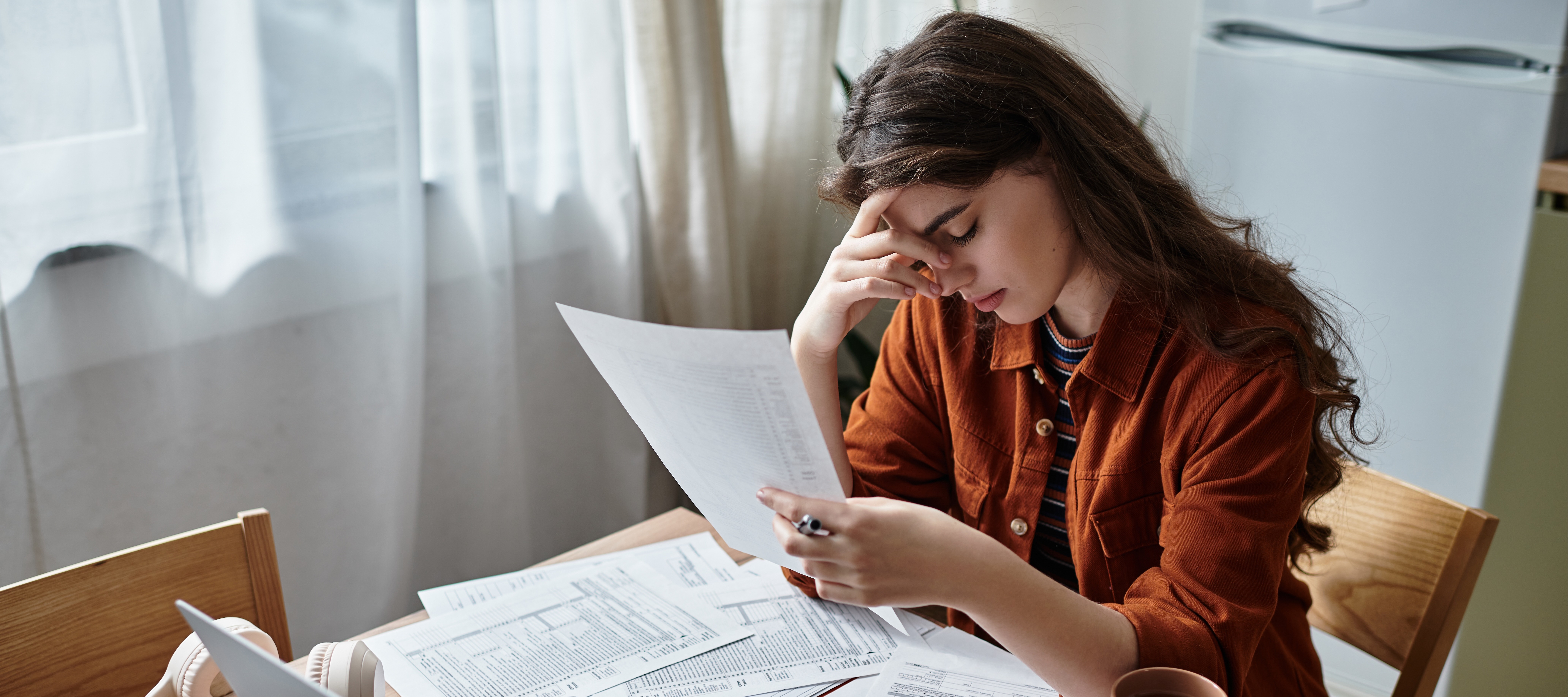 A stressed out looking young woman sits at a wood desk in her home with an open laptop and a pile of papers.