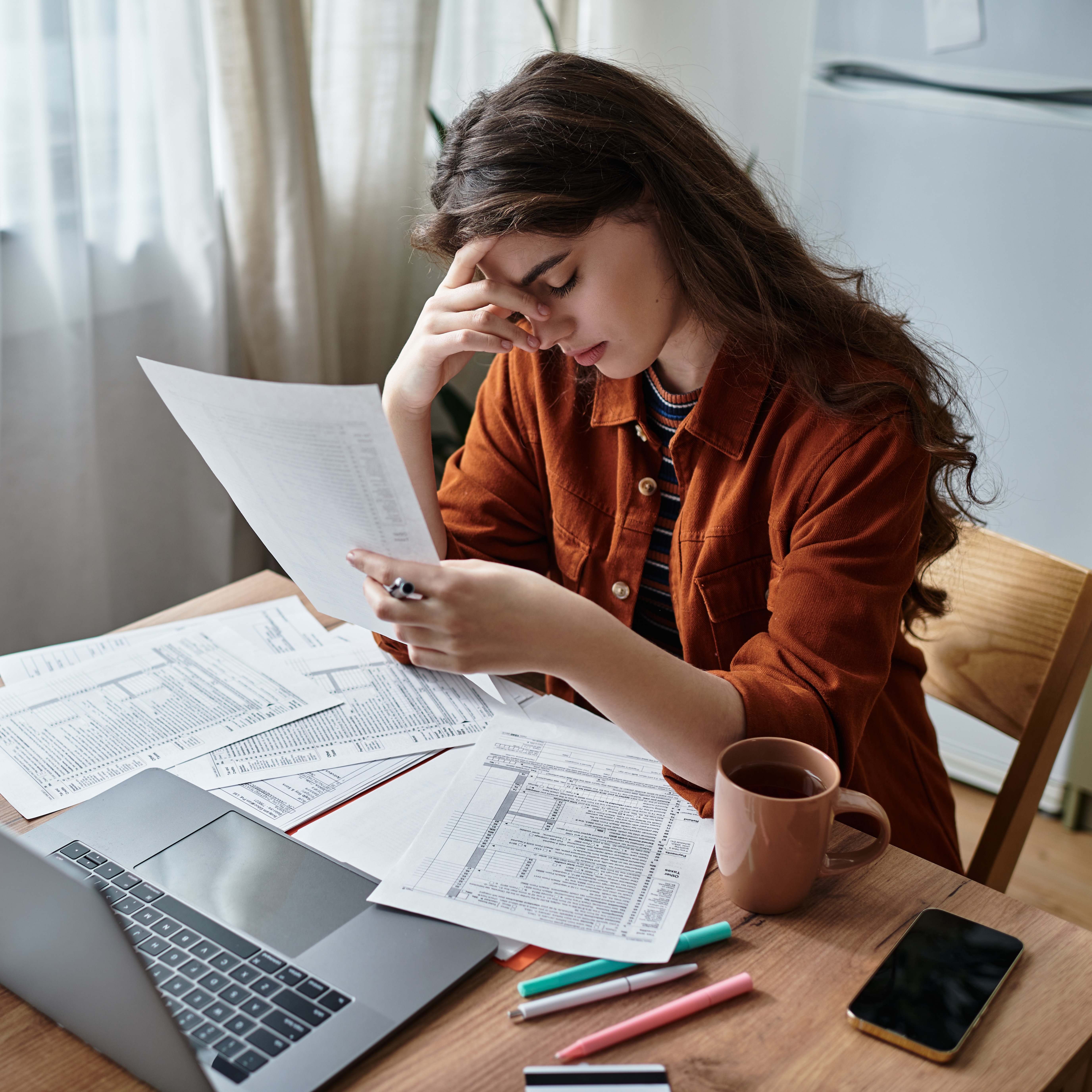 A stressed out looking young woman sits at a wood desk in her home with an open laptop and a pile of papers.